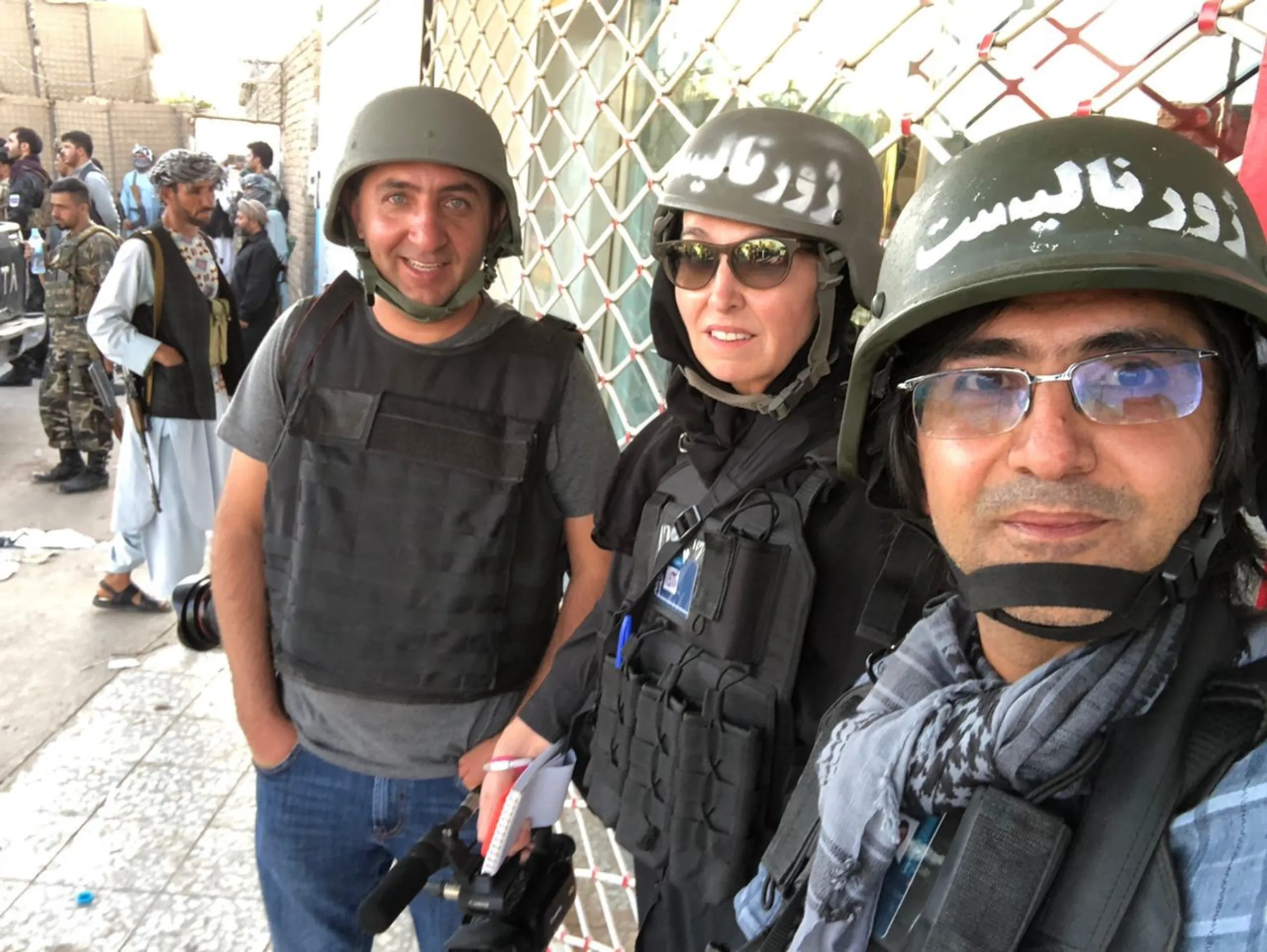 Three people stand in helmets by a chain fence smiling at the camera