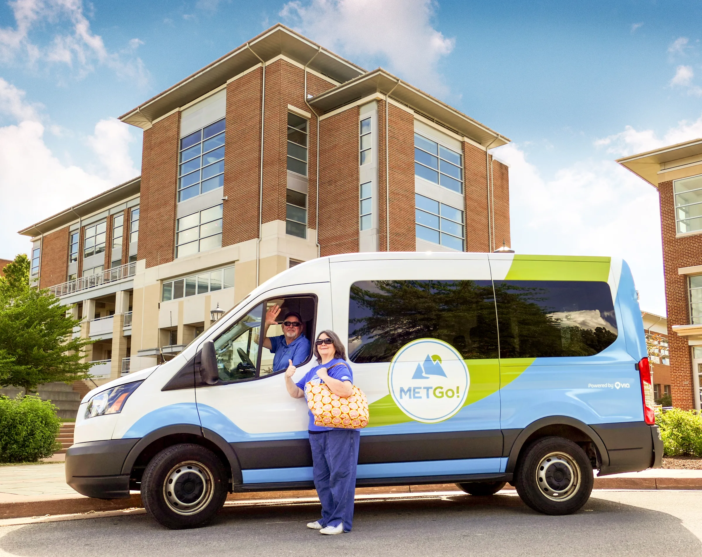 A woman poses in front of a MetGo transit van parked in front of modern buildings, as the driver waves out the window