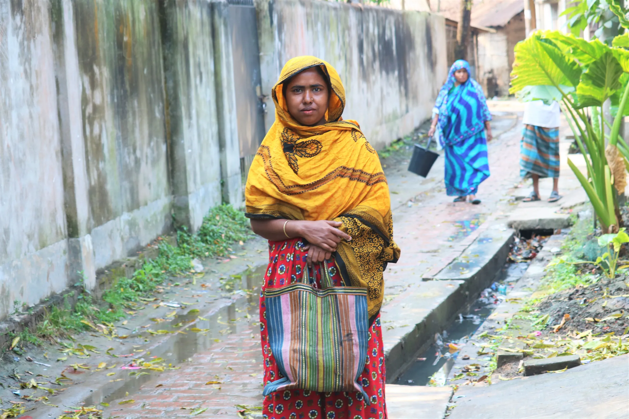 A woman wearing a yellow headscarf stands holding a coloured bag in a street wet from flooding