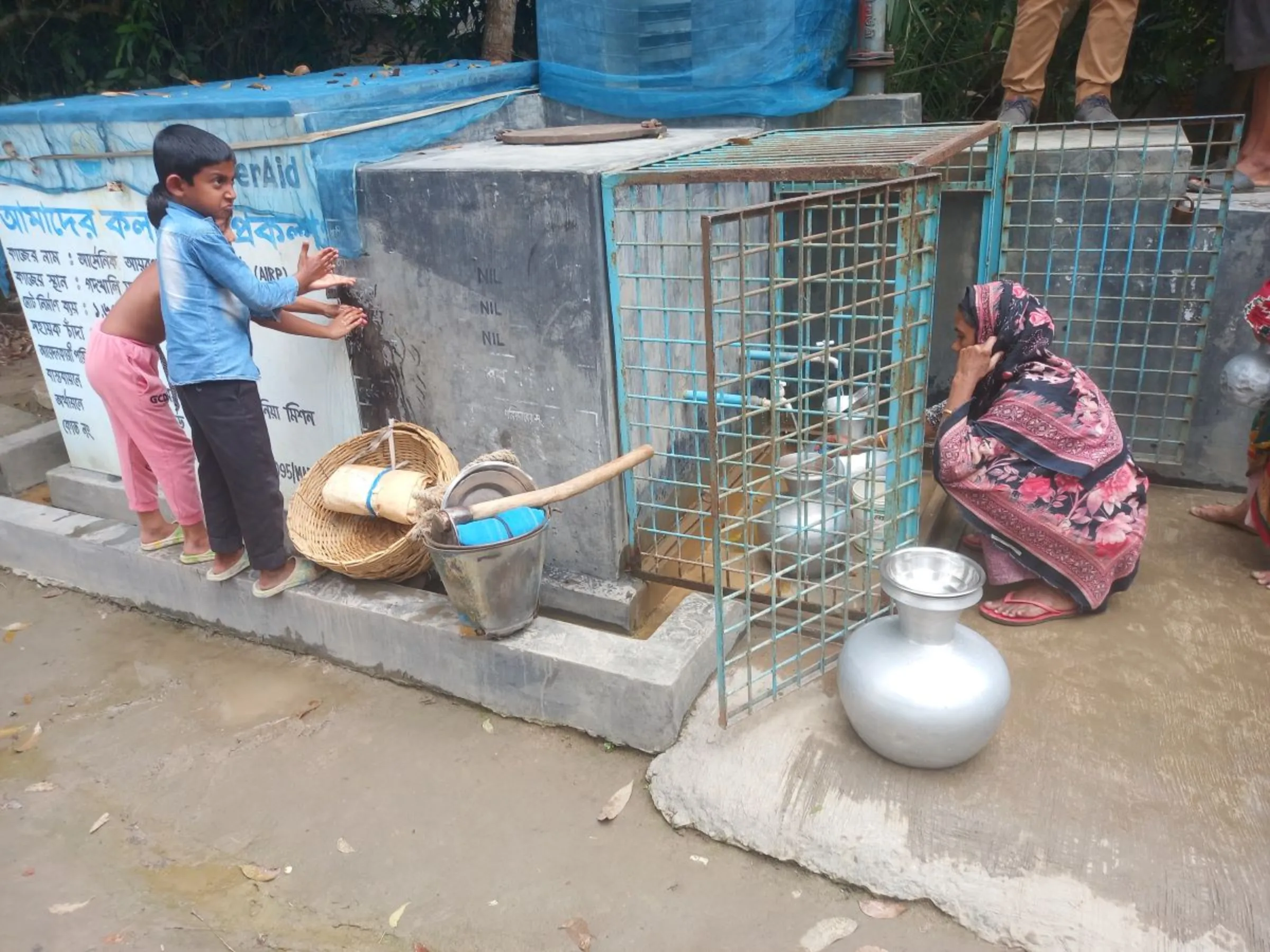 Children stand next to a water facility as a woman crouches to fill up silver pots