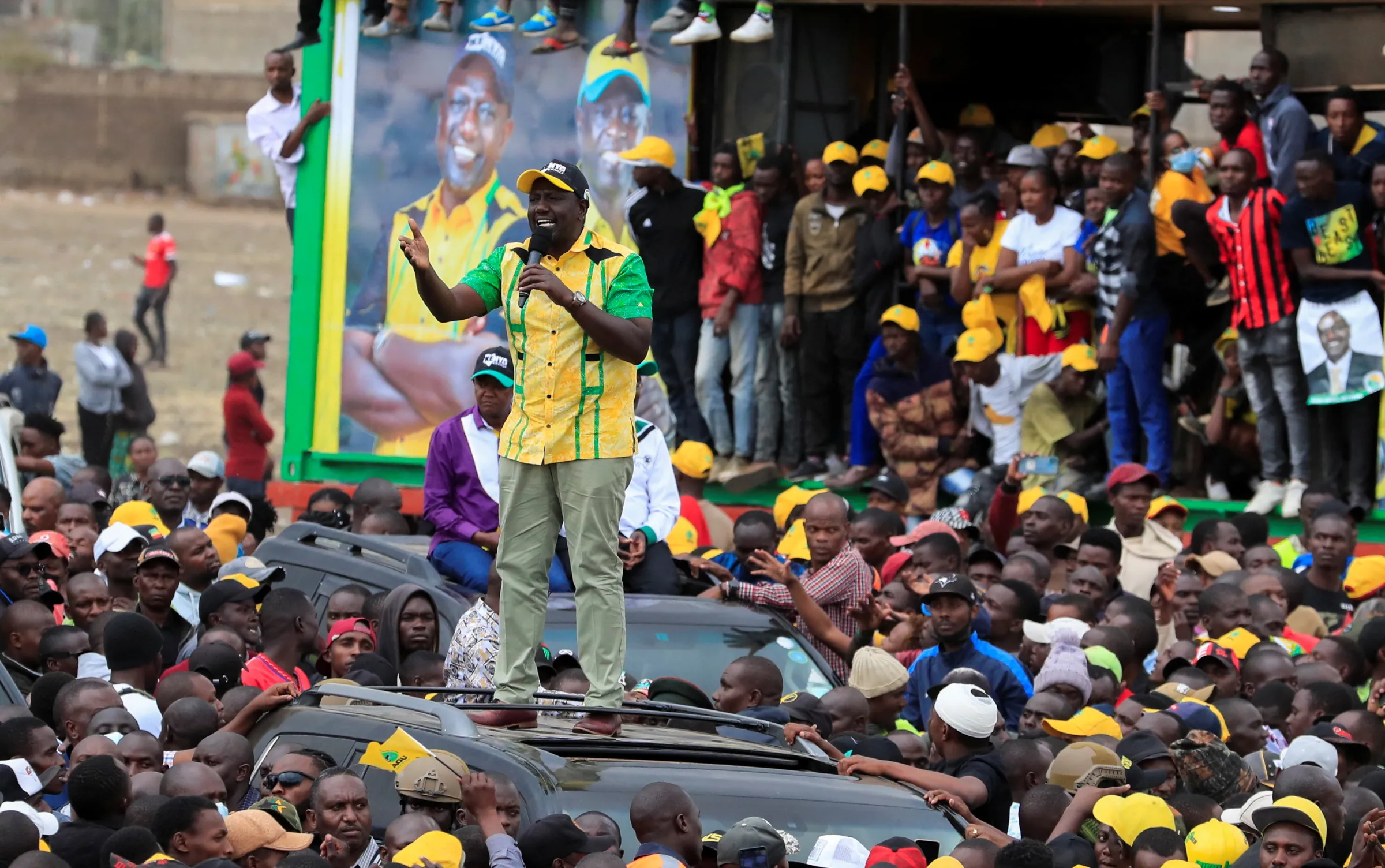 A man stands atop a car roof addressing a crowd with a microphone