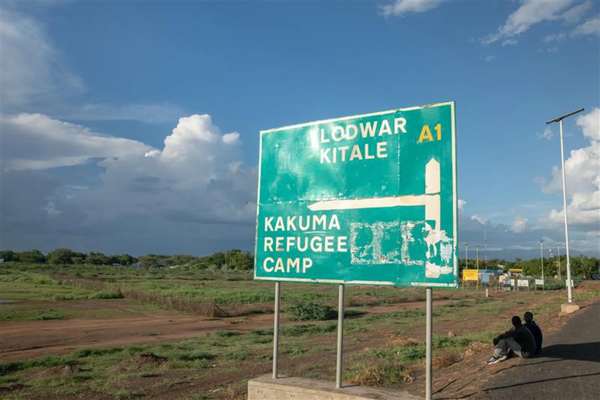 Men sitting by a road sign near Kakuma refugee camp in northern Kenya, May 20, 2025. Thomson Reuters Foundation/Farai Shawn Matiashe