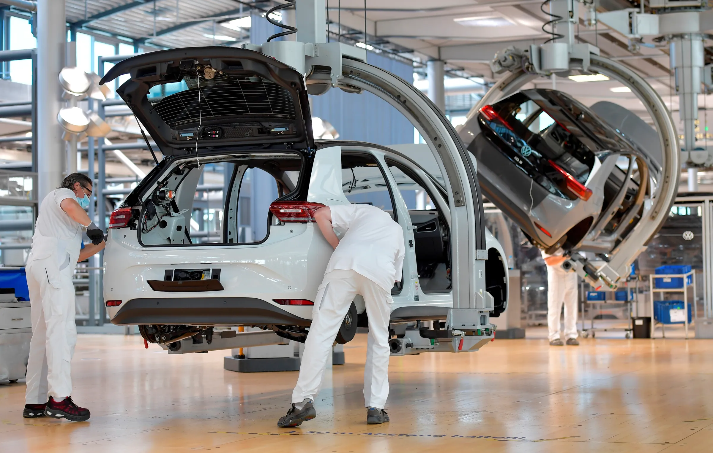 Mechanics in white are working on a car frame which hangs from the ceiling