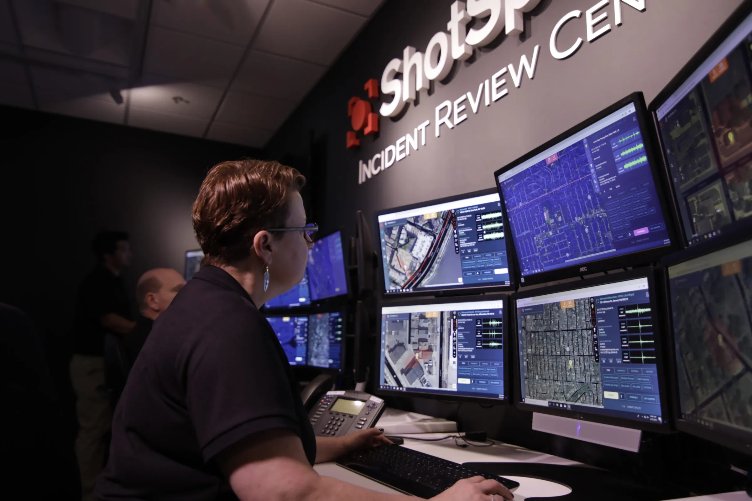 A woman sits at a desk monitoring a computer with several screens