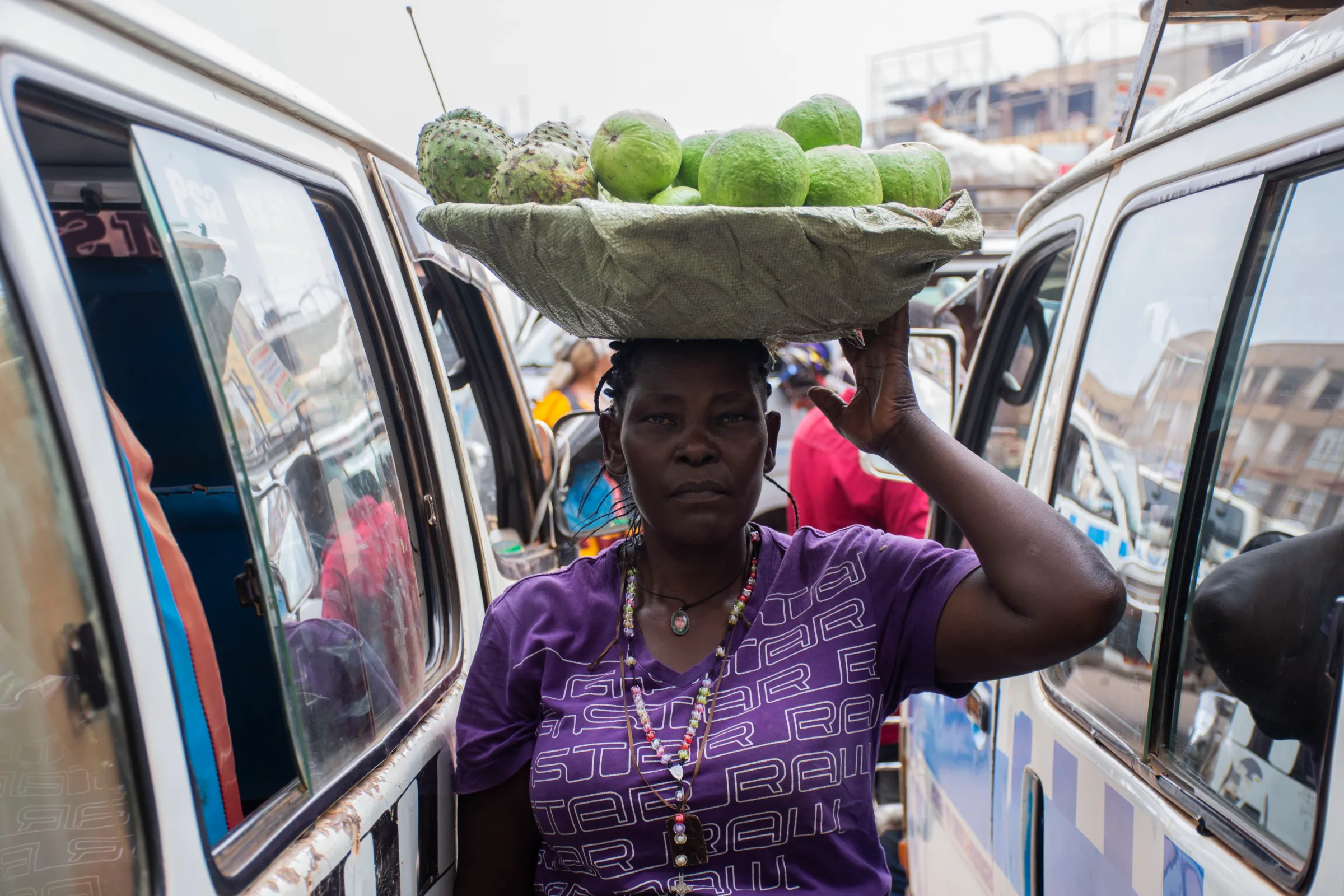 A woman stand between two vehicles, posing for a photo with a basket of fruit resting on her head