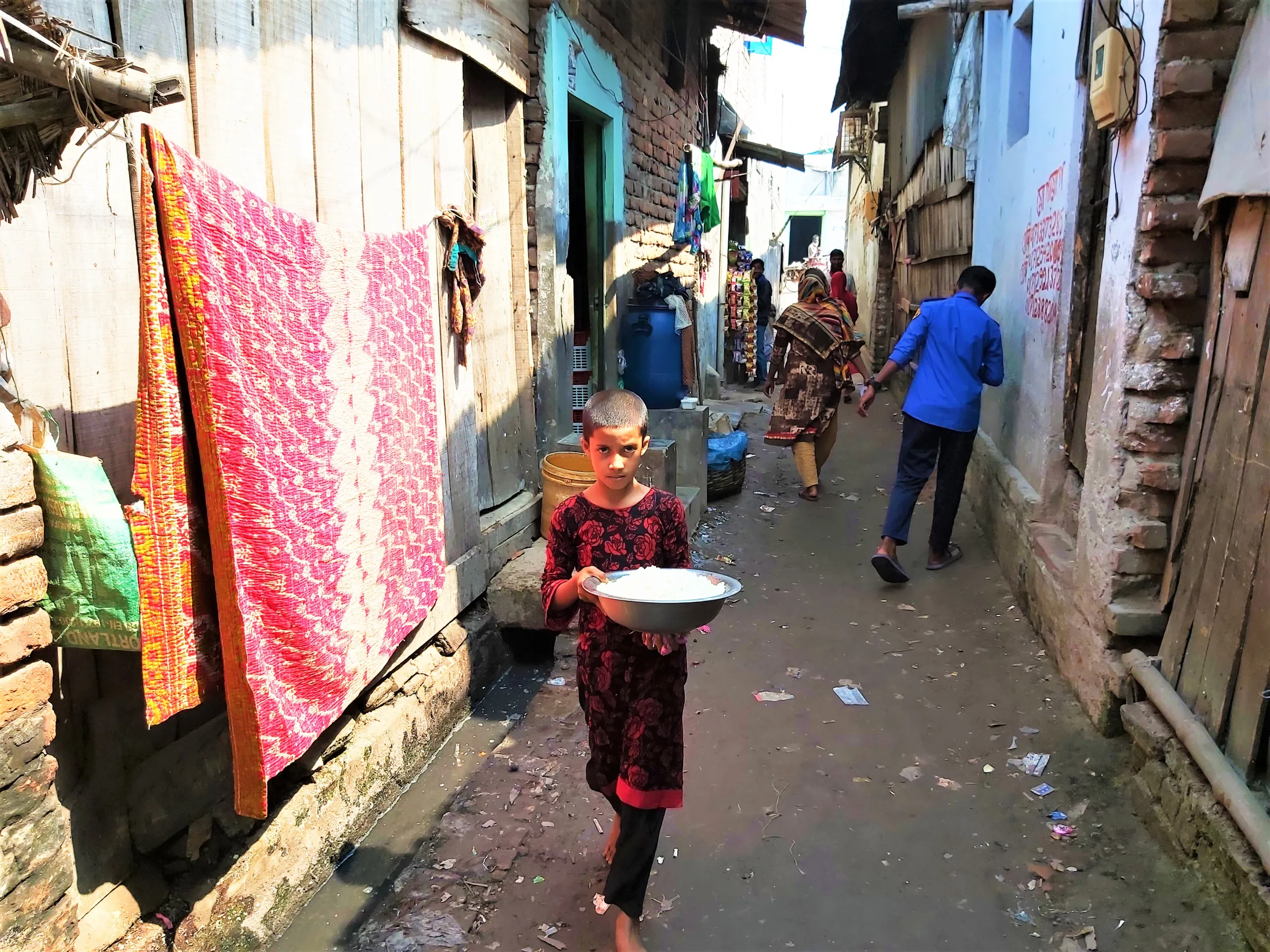A child stands holding a bowl of rice in a narrow street of shacks