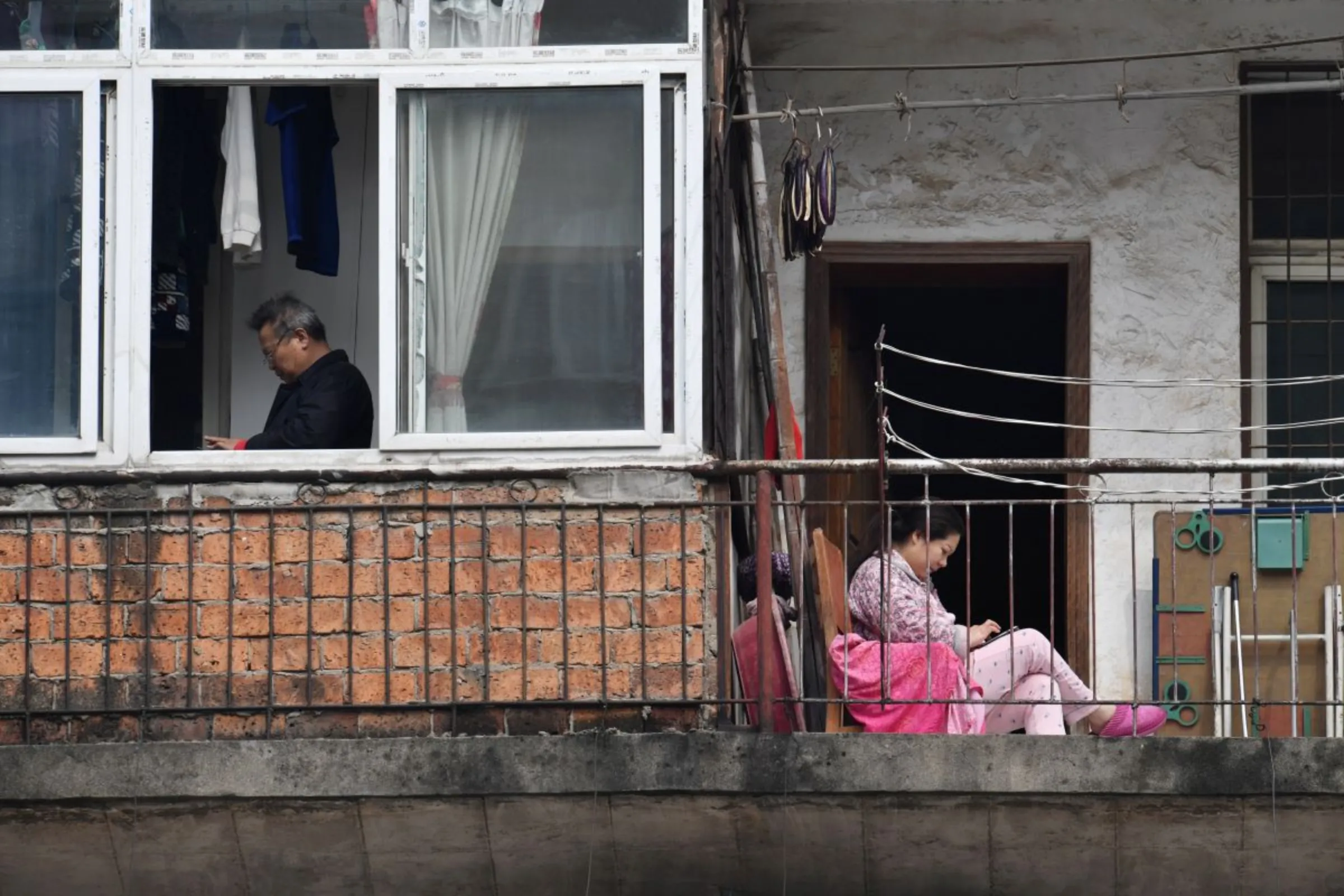 A woman looks at her mobile phone as she sits on a balcony near a man standing by a window at a residential compound in Wuhan, Hubei province, China, March 10, 2020