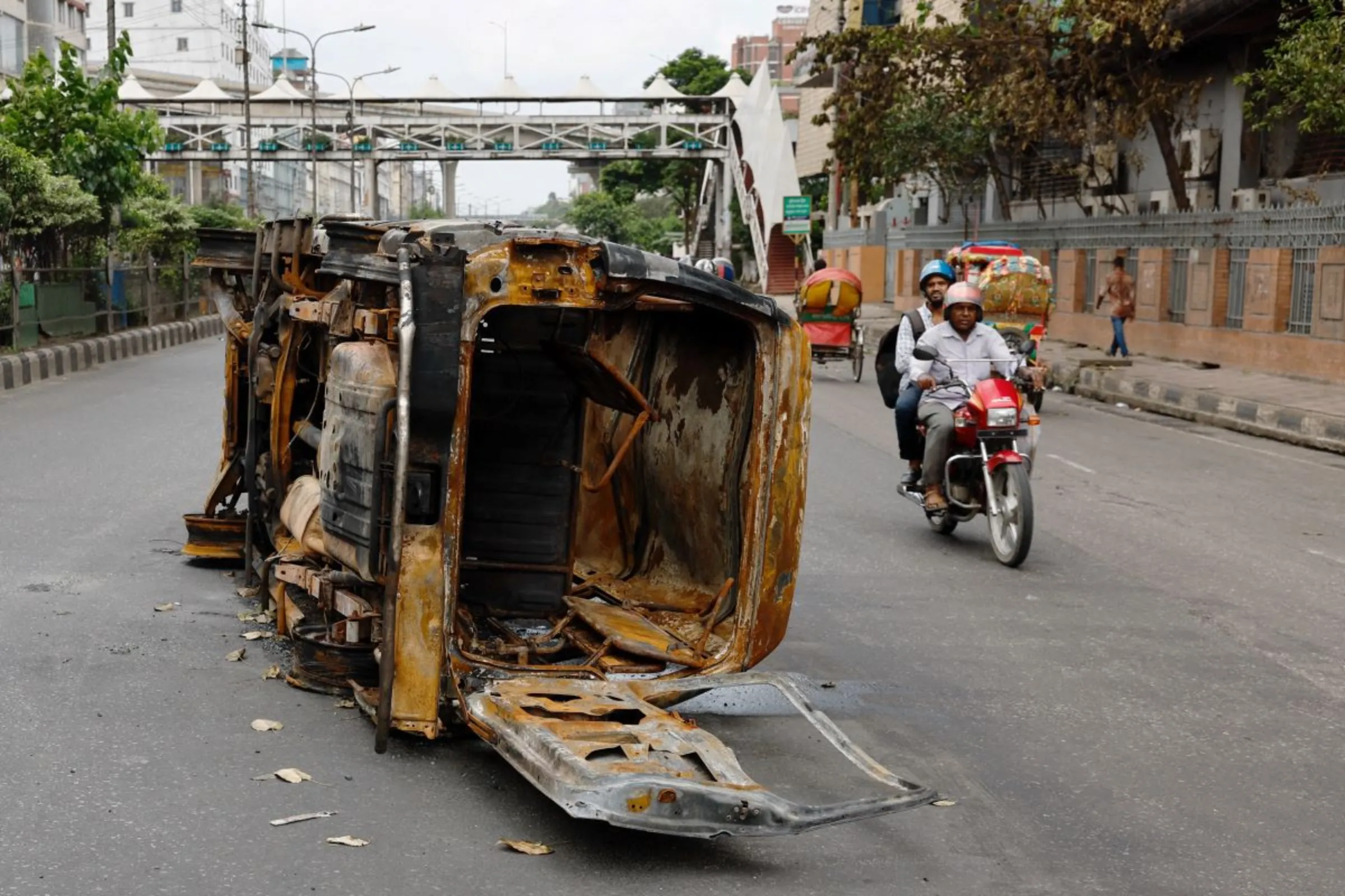 Men ride on a motorbike past a damaged vehicle that was set afire by a mob during clashes after violence erupted following protests by students against government job quotas, in Dhaka, Bangladesh, July 22, 2024. REUTERS/Mohammad Ponir Hossain