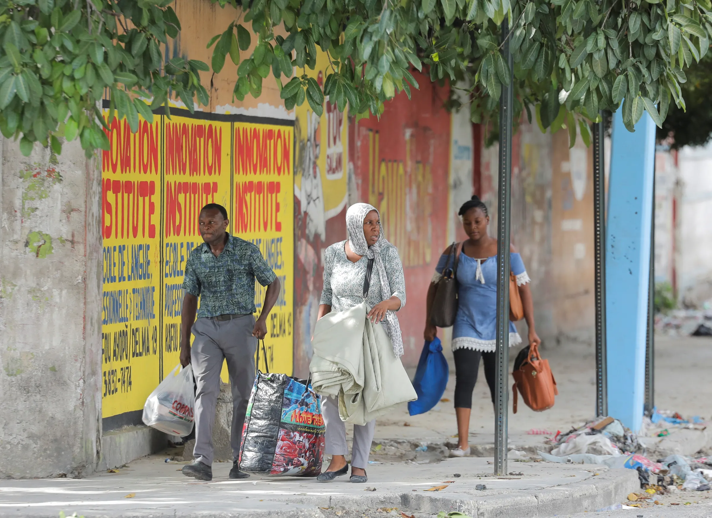 Three people carrying bags walk along a tree lined street