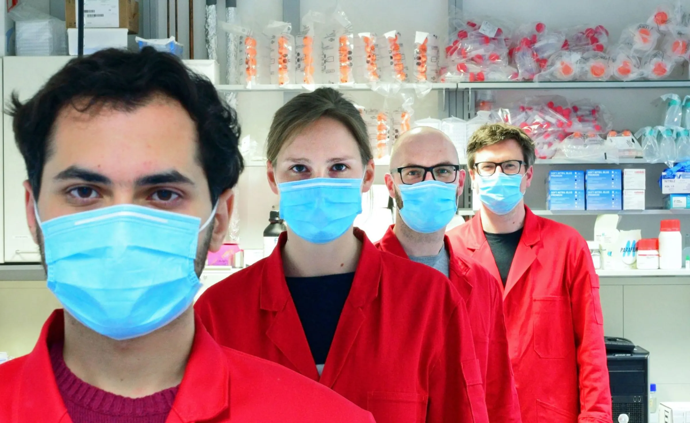 Scientists in red lad coats and protective masks stand in line in a science lab