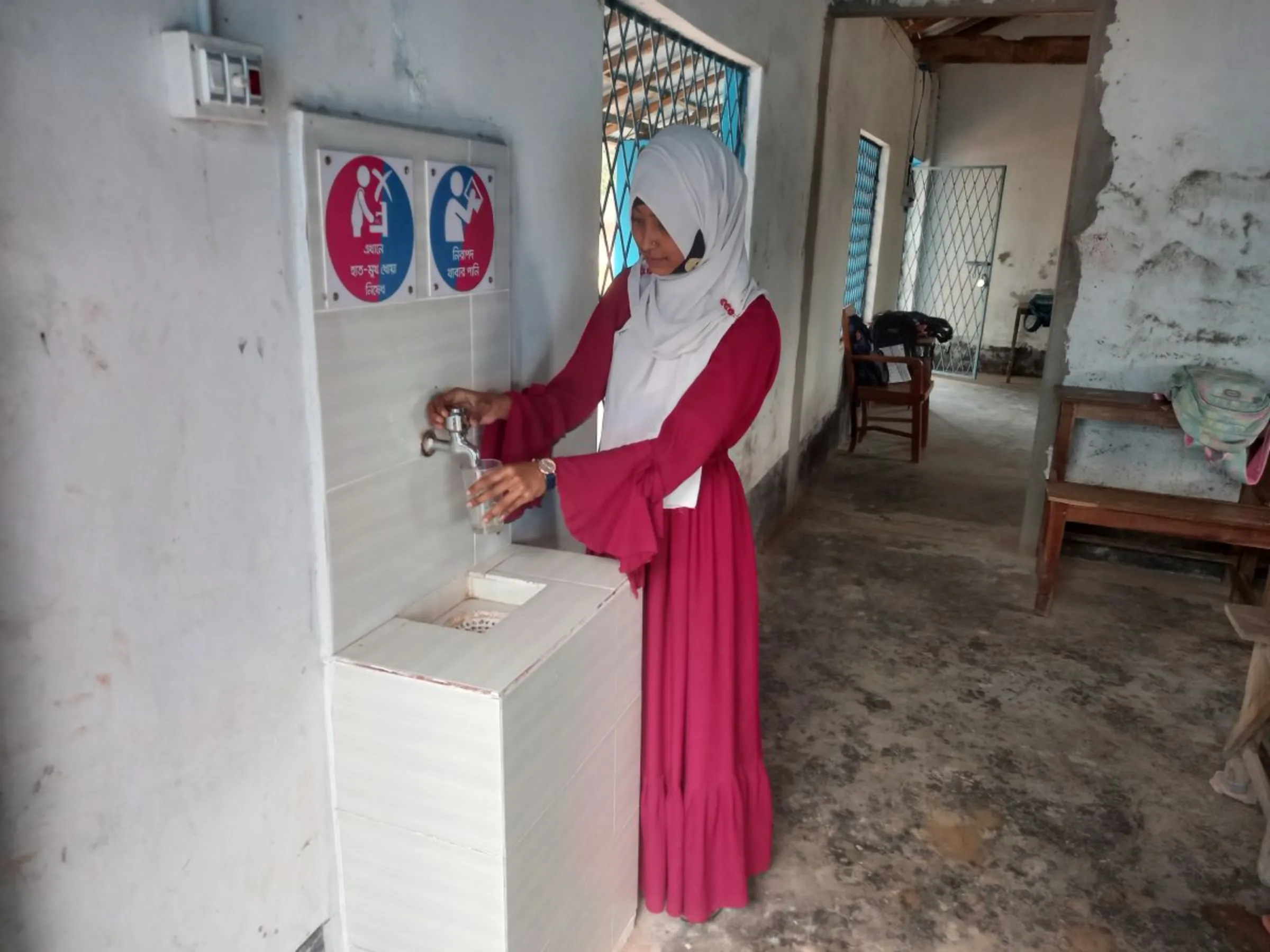 A girl in a pink dress and headscarf stands to pour water from a tap