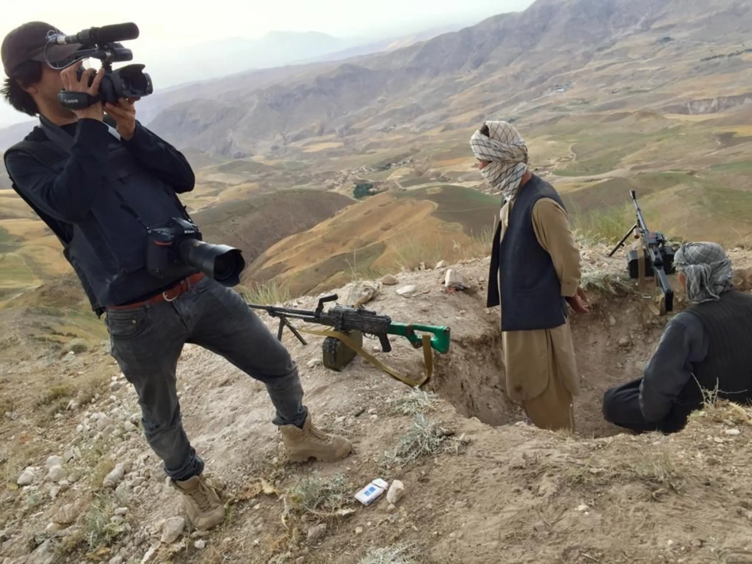 A man films Taliban forces who are watching out from a ditch in the ground, guns propped up
