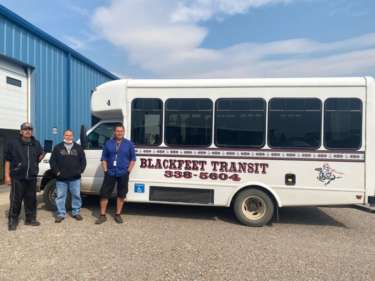 Three men pose next to a Blackfeet Transit minibus parked outside a depot