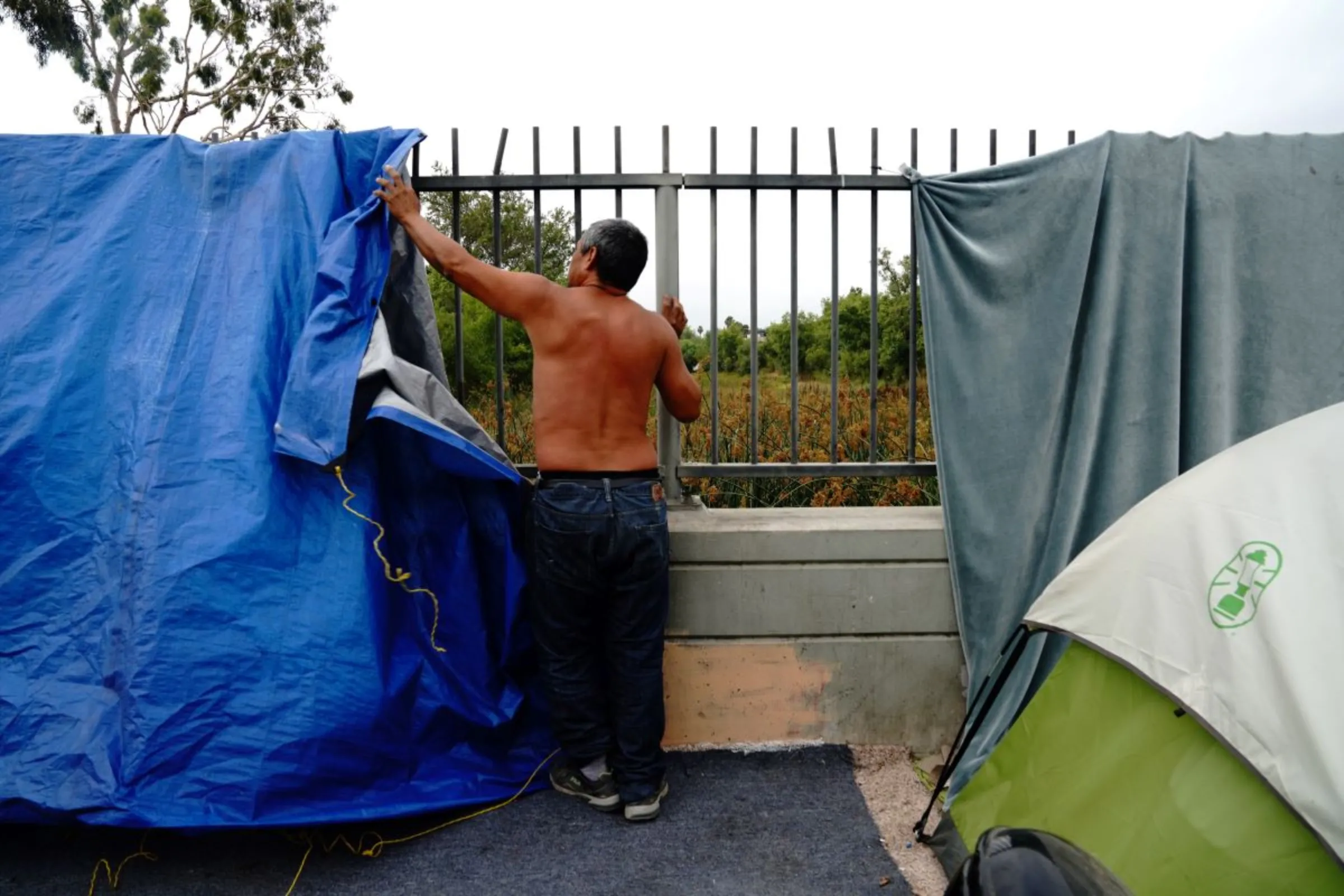 An unhoused man removes tarps and blankets from a fence at a homeless encampment, before the encampment's clearing by Los Angeles police and sanitation workers in Harbor City, Los Angeles, California, U.S., July 1, 2021. REUTERS/Bing Guan