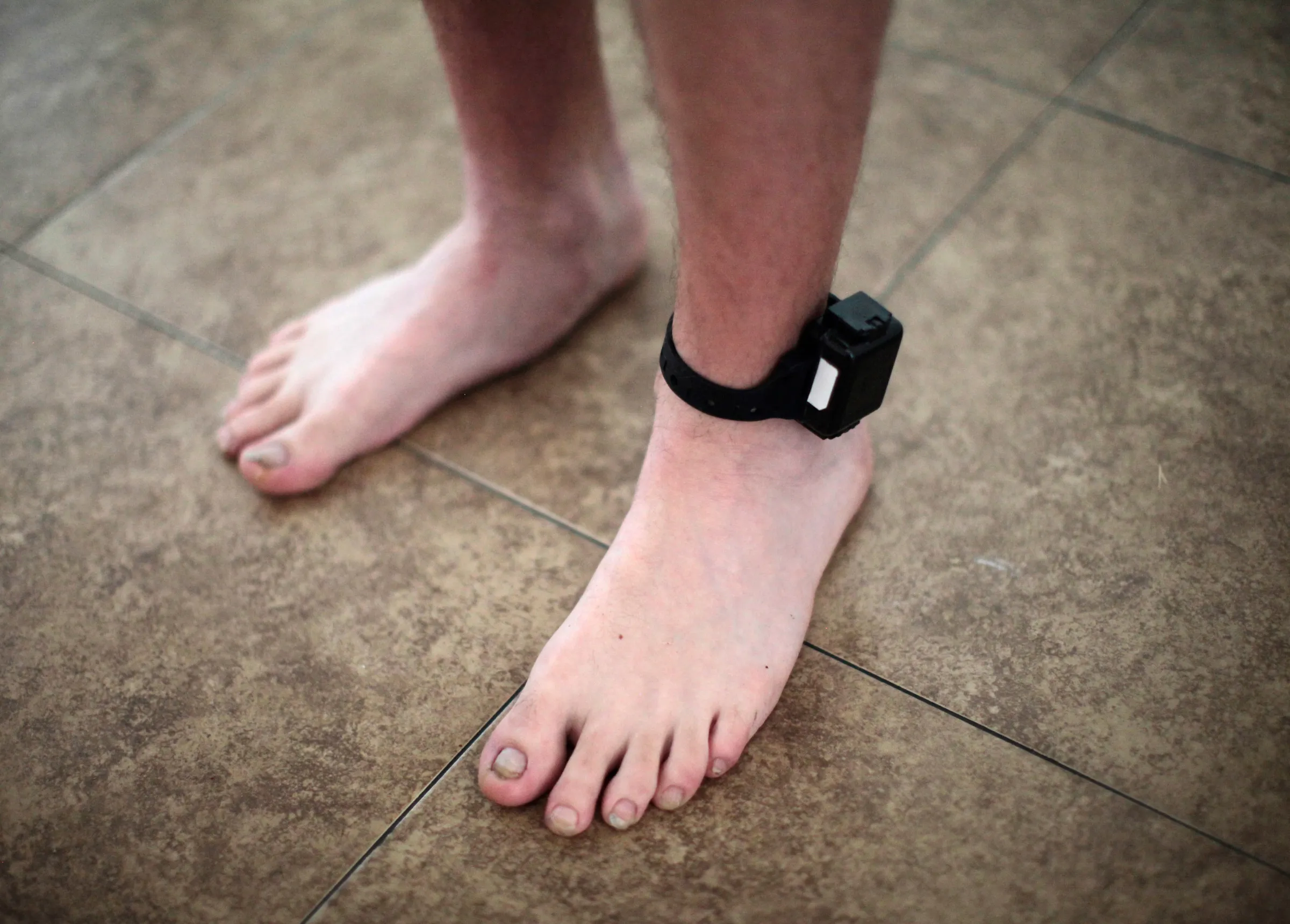 A person's feet are pictured standing on paving stone with an electronic tag around one ankle