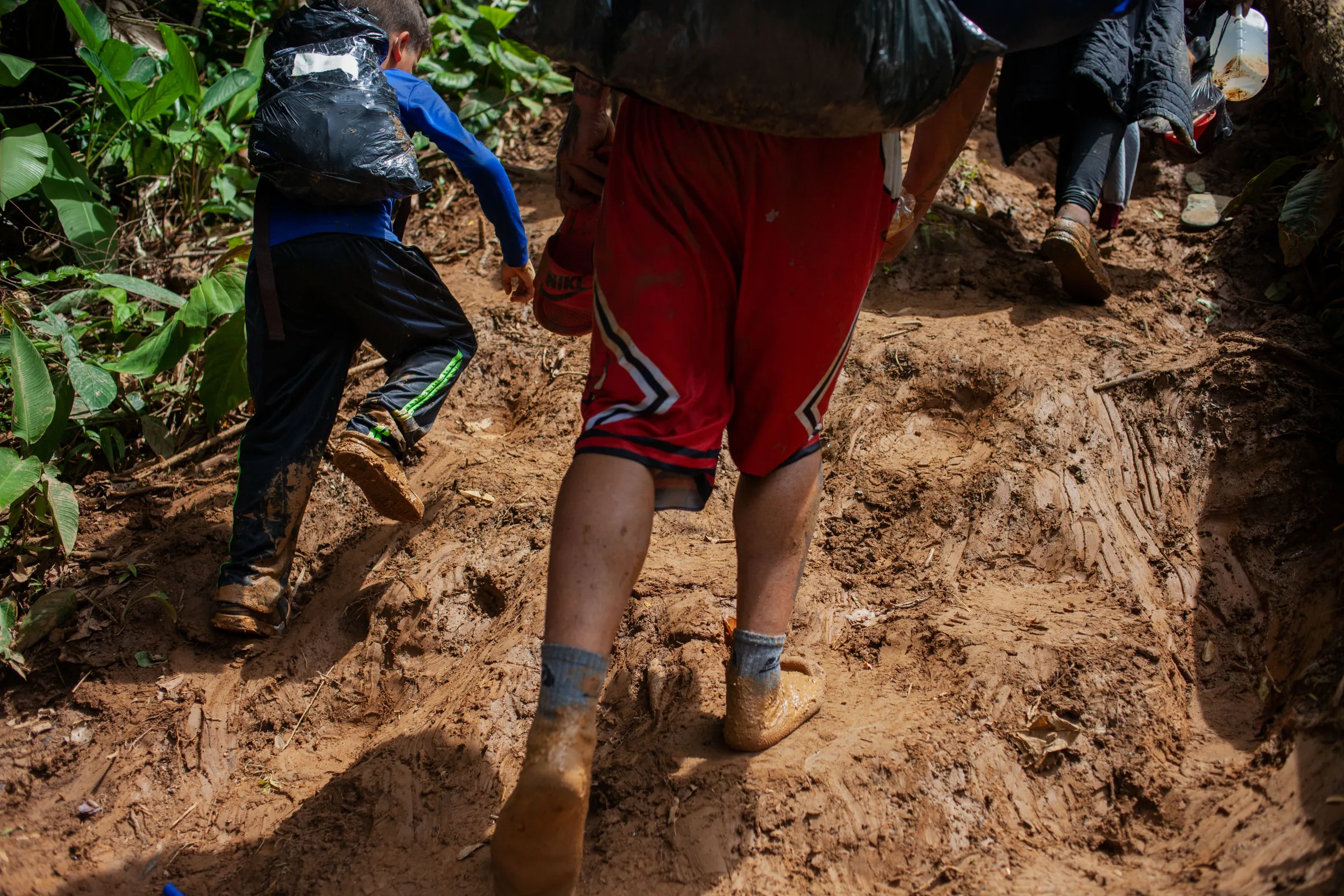 A Venezuelan migrant walks in socks alongside a child as they walk along a muddy path through the Colombian jungle bordering neighboring Panama at the start of a seven-day trek. Darién Gap, Colombia, July 27, 2022. Thomson Reuters Foundation/Fabio Cuttica