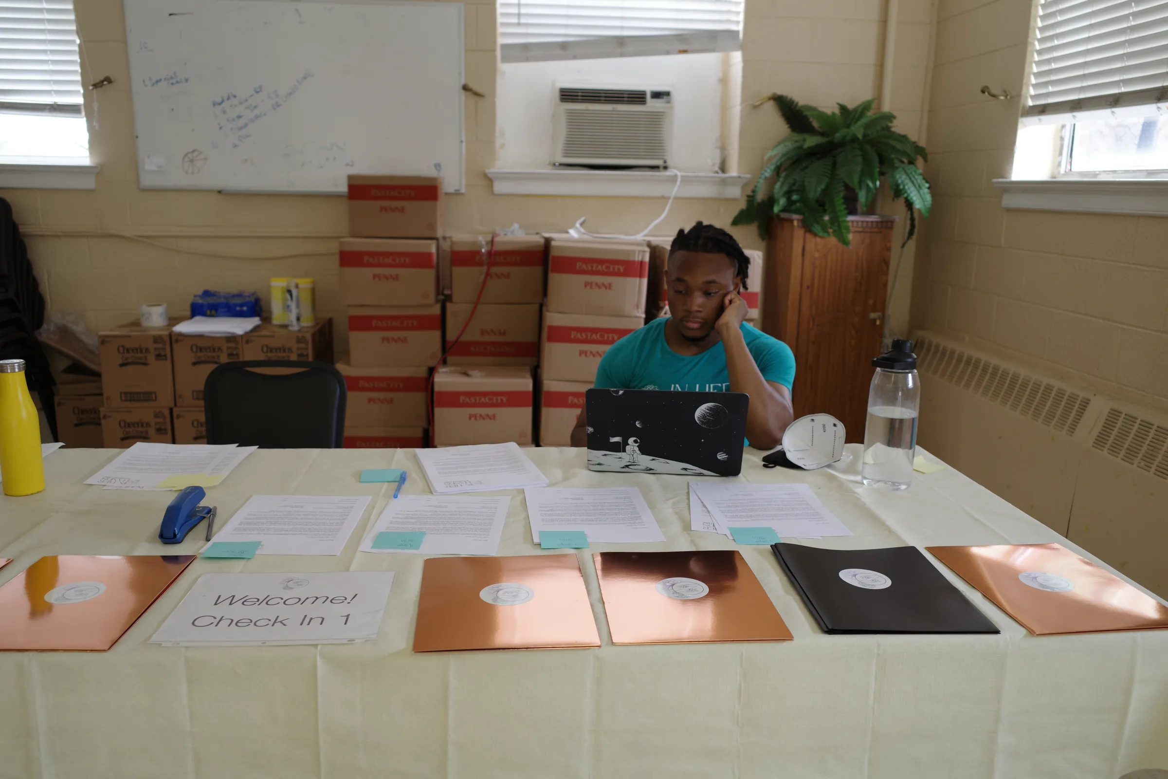 A man sits working on a laptop at a desk covered in piles of paper, stacked boxes line the walls behind