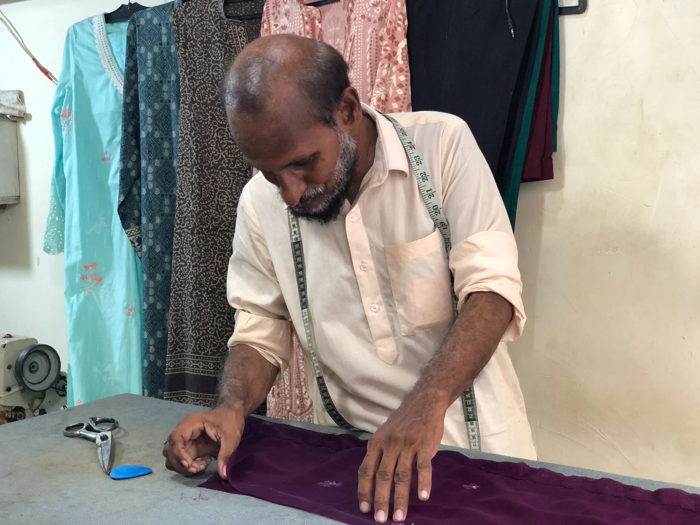 A tailor measures a piece of cloth on a bench, with dresses hanging on the wall behind him