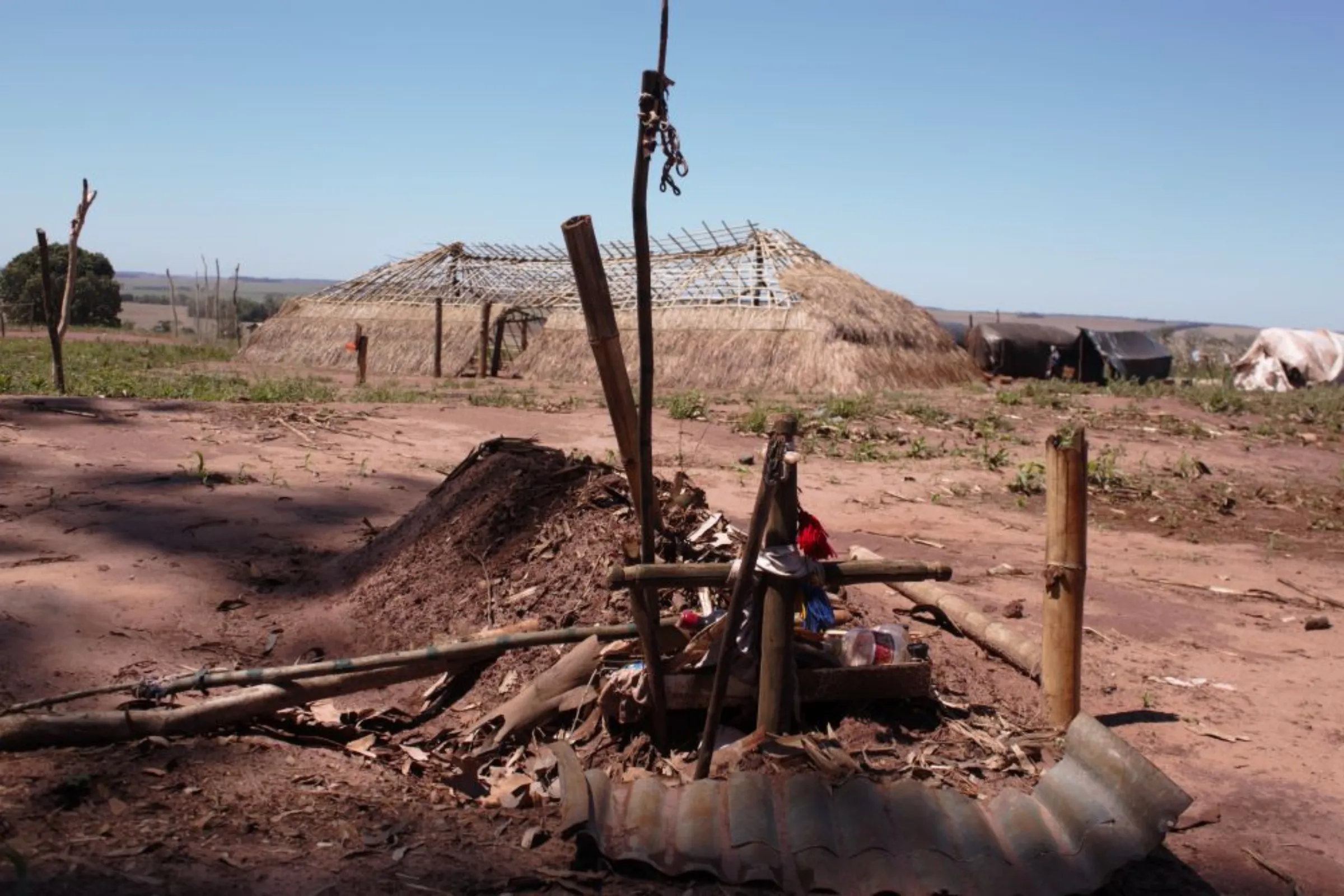 Grave of Vitor Fernandes, a Guarani-Kaiowá prayer-man killed during a Military Police raid in the 24th of June; at the backdrop a prayer house (“oga pysy” in Guarani) under construction near barracks in the Guapoy occupation, by the Amambai Indigenous Reserve, in Amambai, Brazil, September, 17, 2022. Thomson Reuters Foundation/André Cabette Fábio