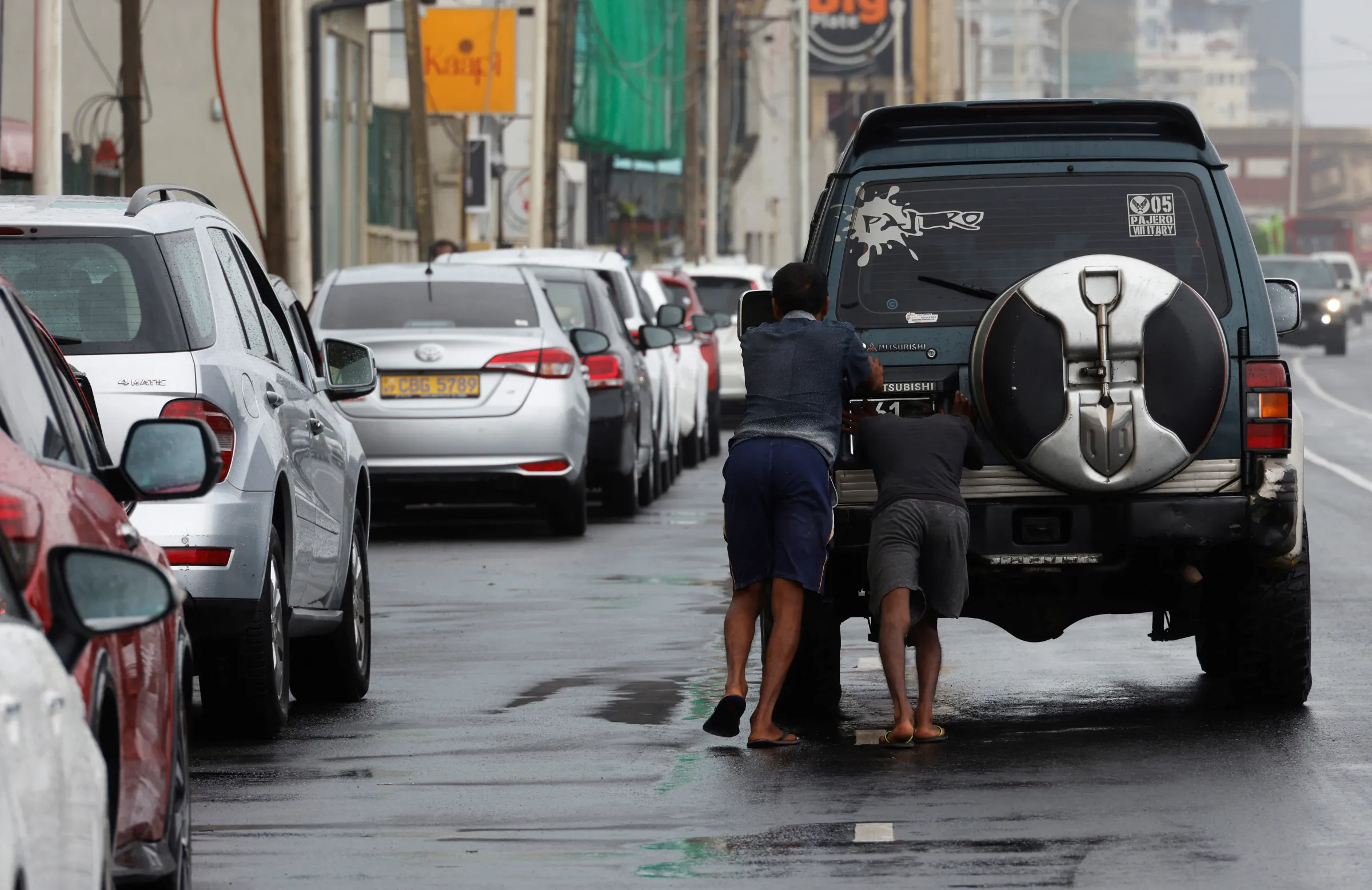 Men push a car as its battery is discharged next to a line where cars wait to buy petrol from a fuel station
