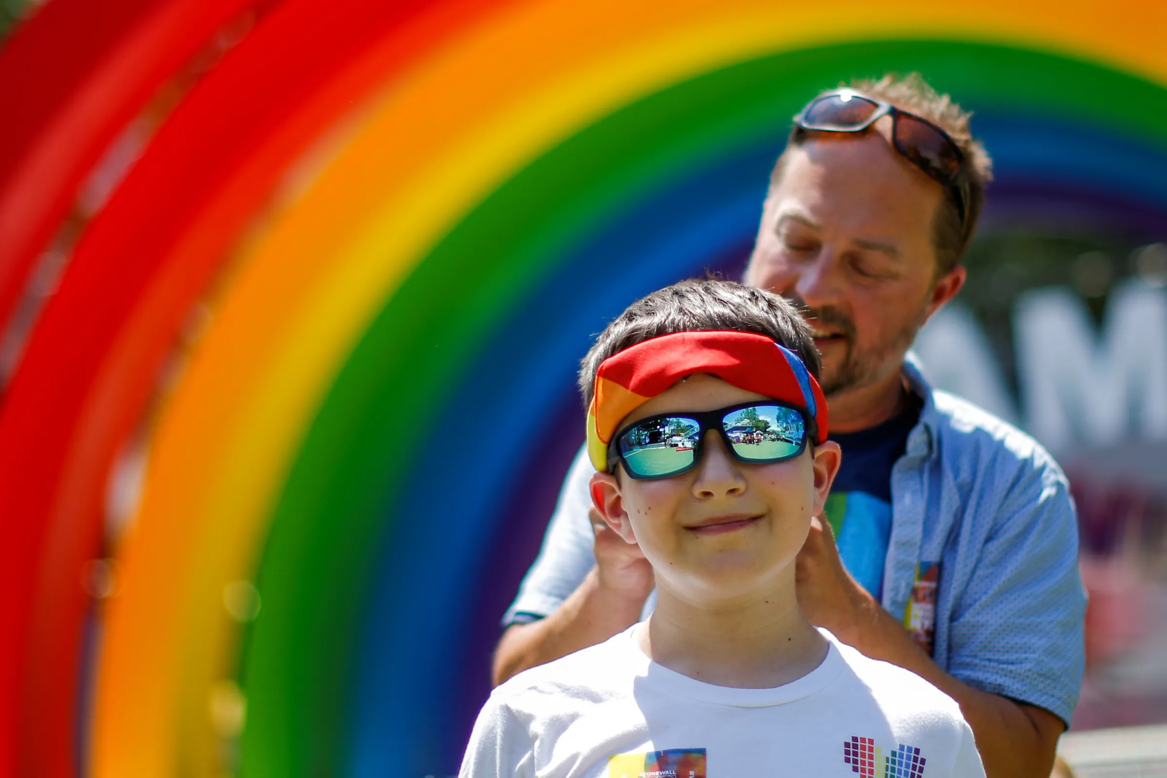 A man ties a rainbow headscarf around a boy as they stand in front of a giant rainbow