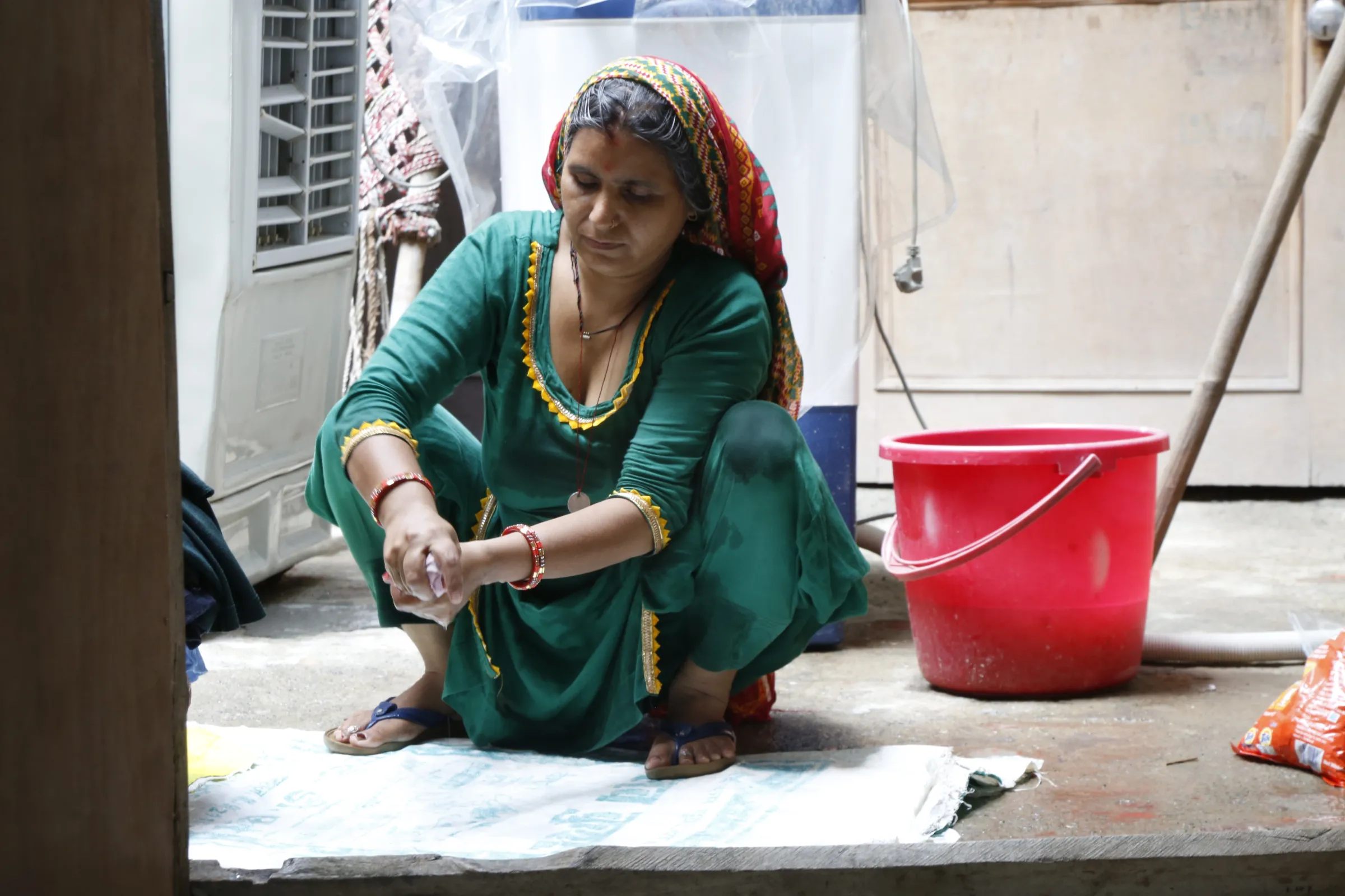 A woman, who suffered two miscarriages over the last year in an attempt to have a son, squats beside a red bucket washing clothes in her village home in Hisar, India, July 31, 2022. Thomson Reuters Foundation/Annie Banerji