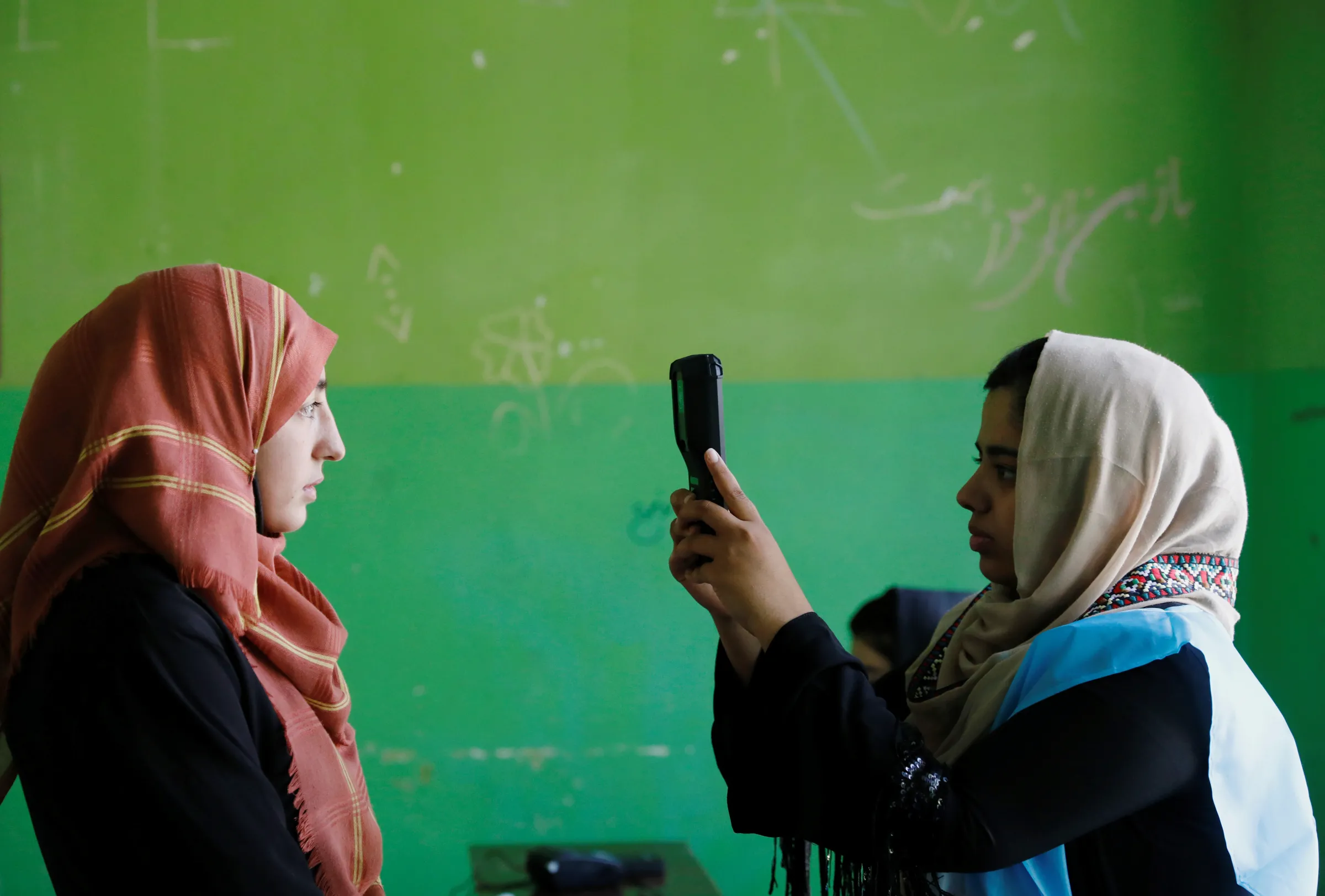 A woman holds up a device to take a picture of someone standing in front of a green wall