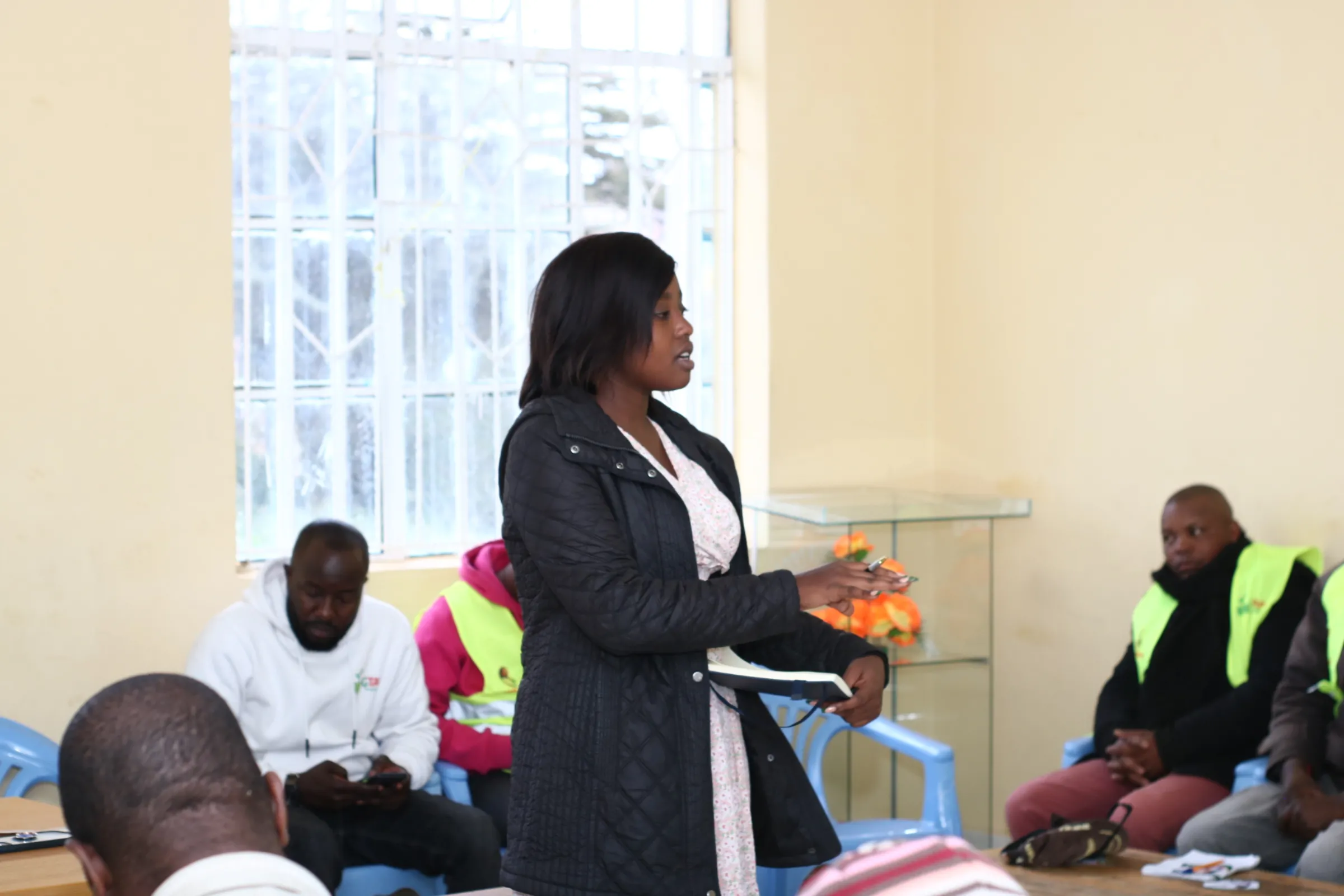 A woman stands speaking to a group of men who are sitting in blue plastic chairs around her