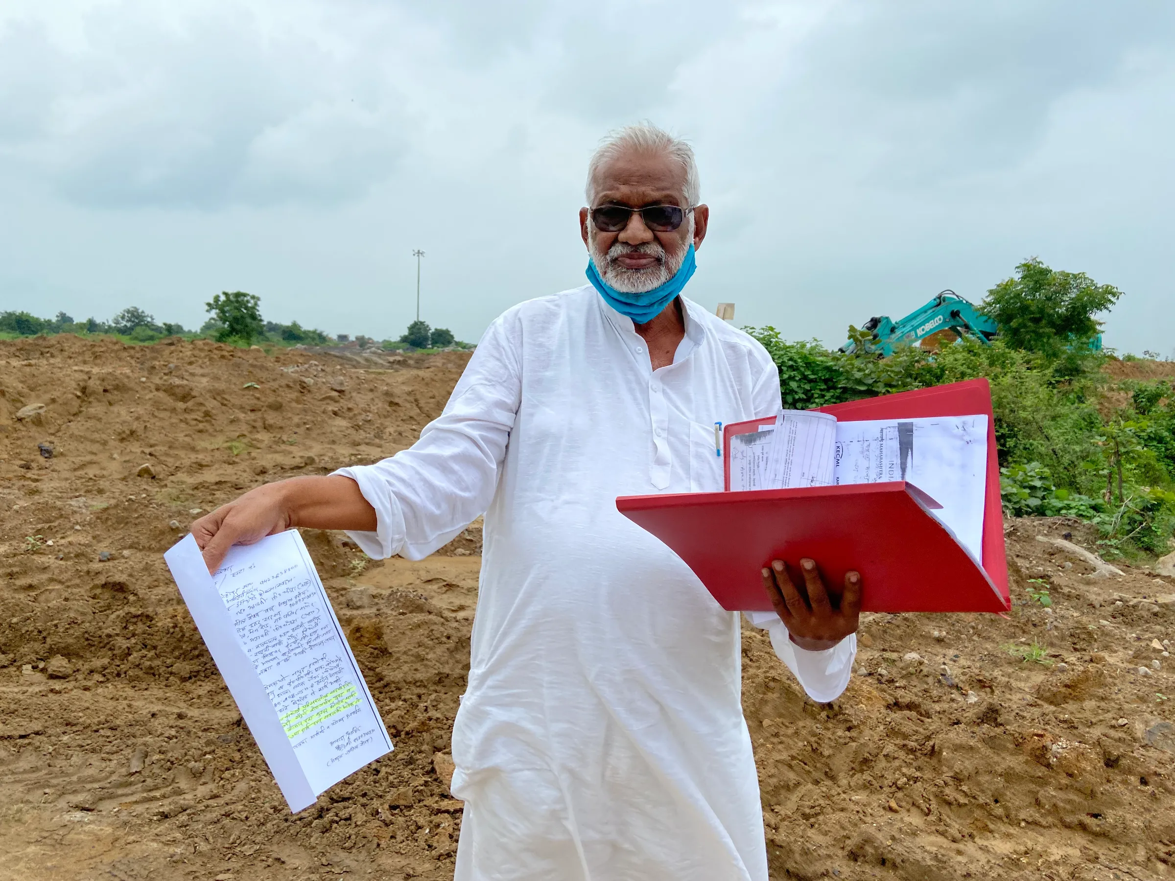 A man wearing sunglasses stands holding documents and a red file in front of a mound