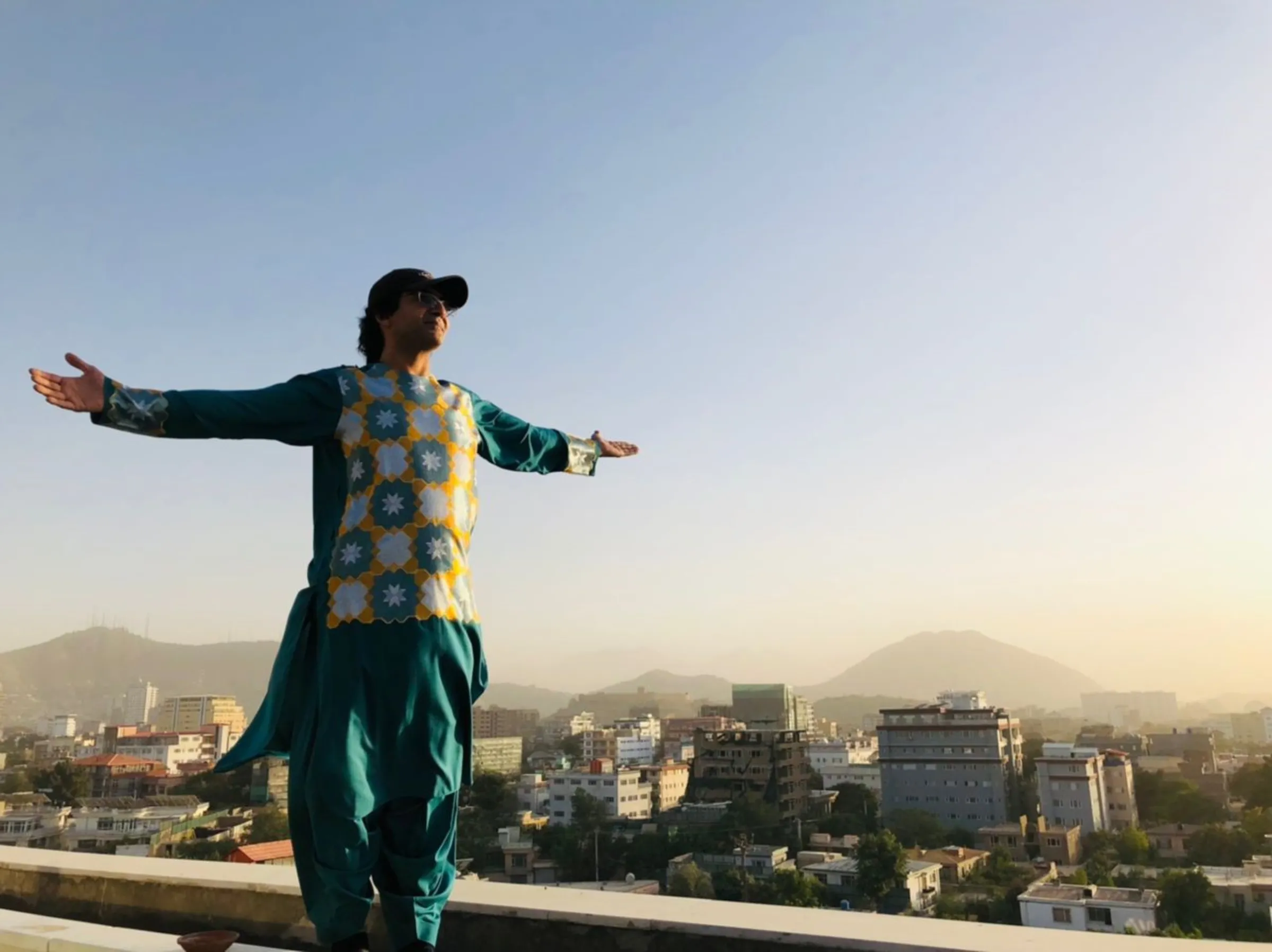 A man stands with his arms outstretched on a Kabul rooftop wearing a traditional Herati outfit