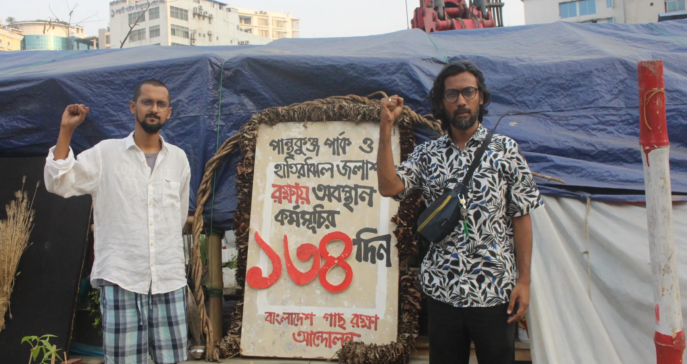 Amirul Rajiv and Naim Ul Hasan, two coordinators of Bangladesh Tree Protection Movement, pose before the makeshift tent where they have been staging a sit-in for more than four months, Dhaka, Bangladesh, April 26, 2025. Md. Tahmid Zami/ Thomson Reuters Foundation