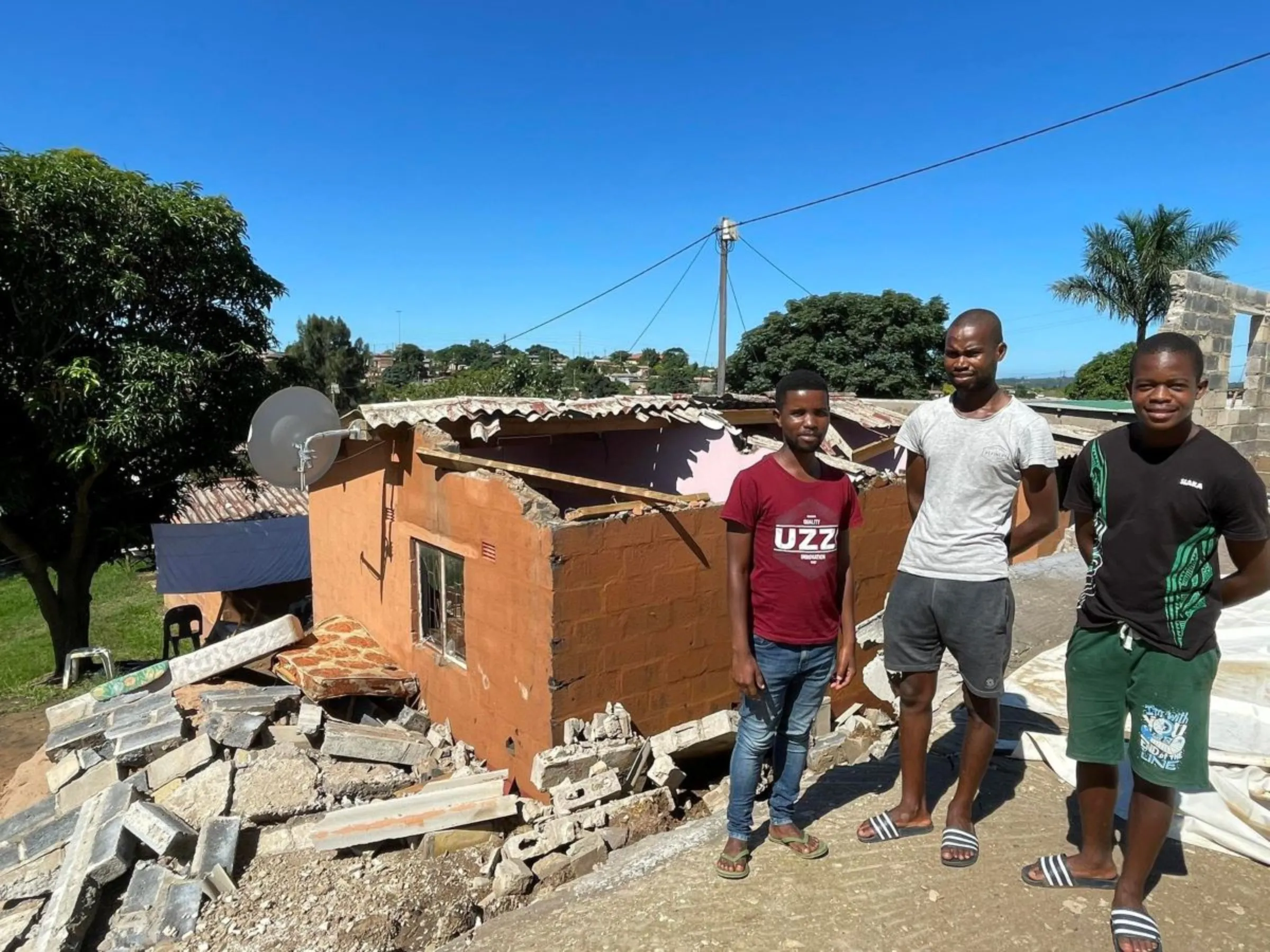Ndumiso Mkhwanazi (L), Siya Sumede and Simanga Shandu (R) stand outside their home without a roof after a church wall collapsed on it during the rains in Shakaskraal township in Durban, April 14, 2022. Thomson Reuters Foundation/Kim Harrisberg