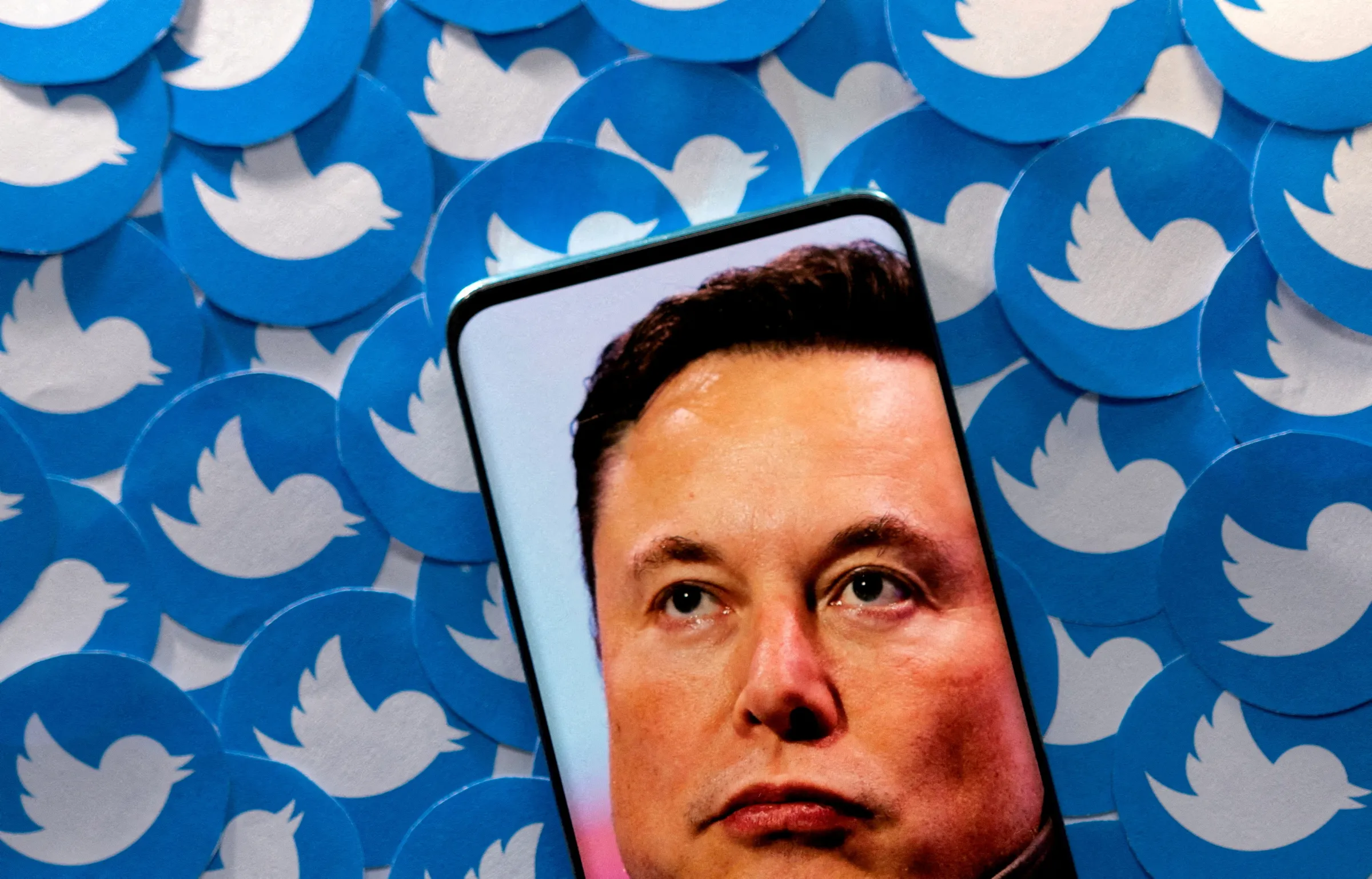 An image of Elon Musk is seen on smartphone placed on printed Twitter logos