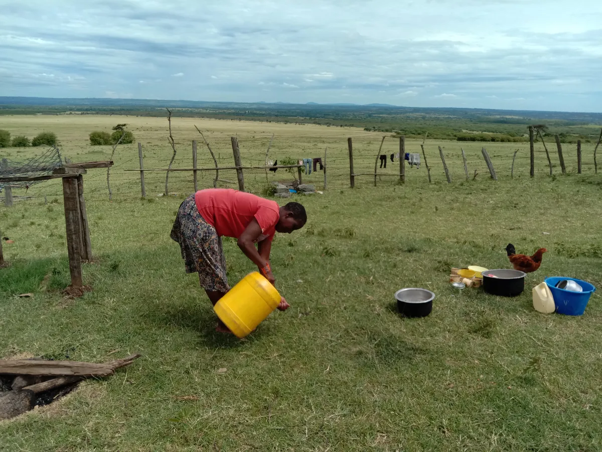 A woman bends over the grass washing dishes with a yellow bucket