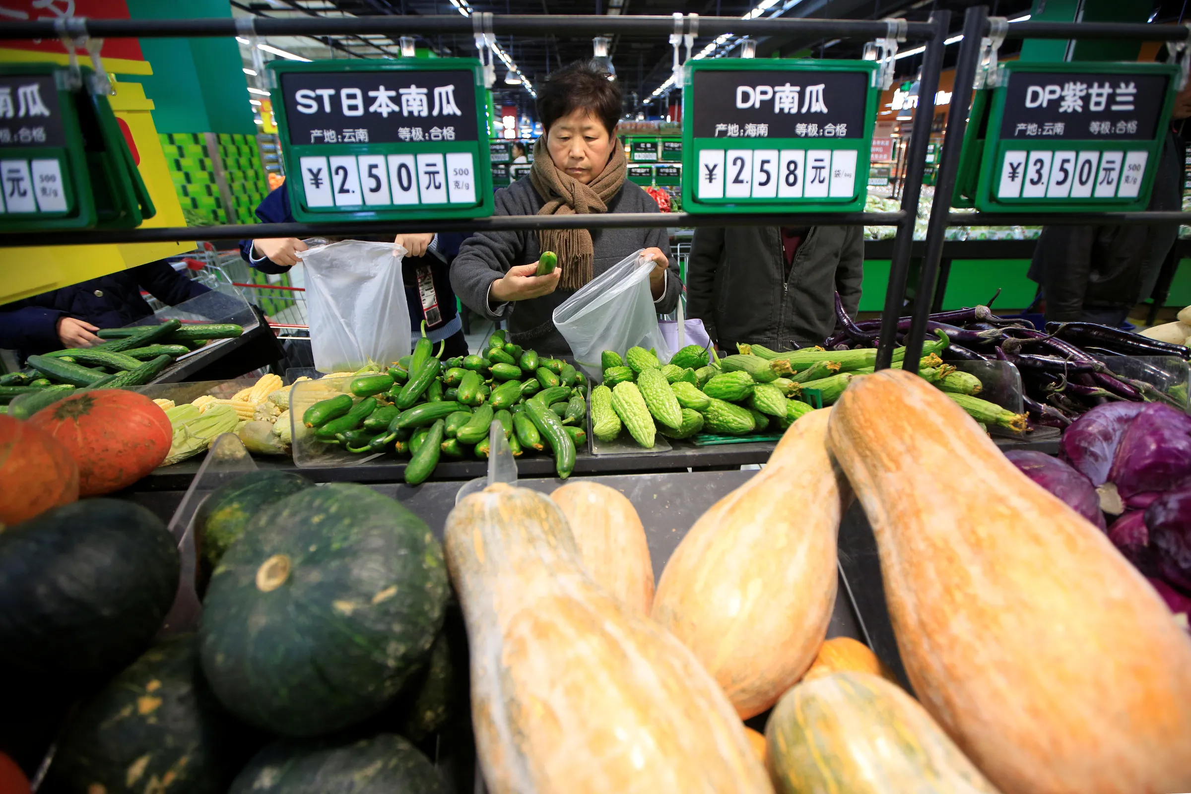 A woman stands holding a plastic bag and a courgette at a vegetable stand