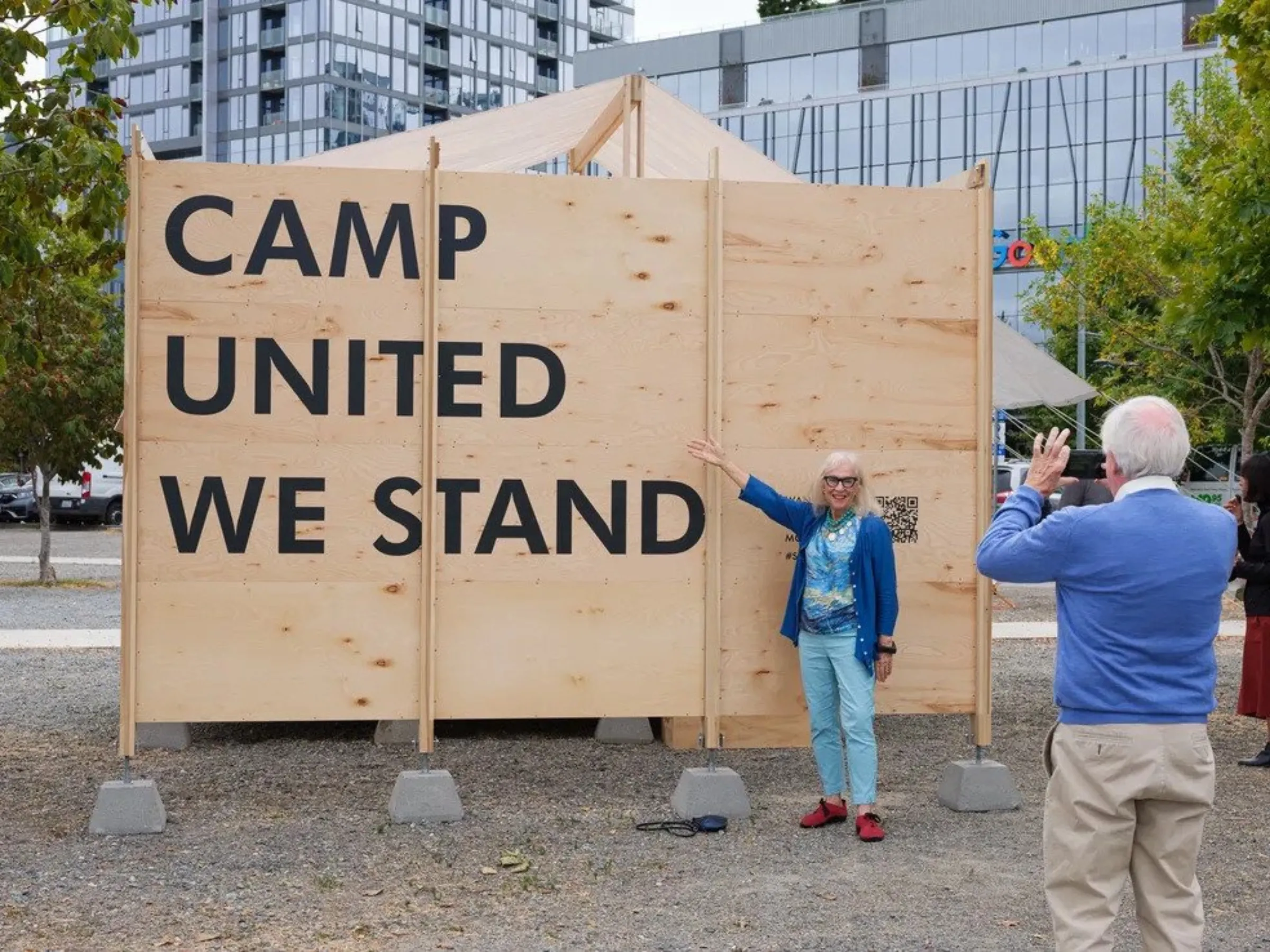 A man takes a photograph of a woman stood in front of a large wooden placard