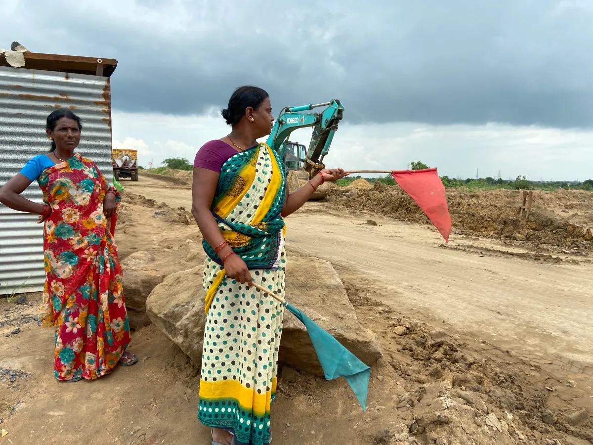 A woman holding red and green flags stand by the side of a mud road, another woman stands behind her