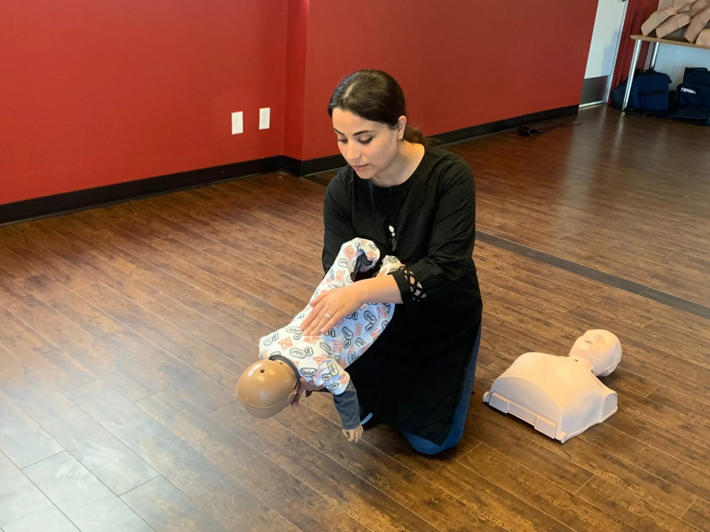 A woman kneels on wooden flooring as she handles a dummy child she is using for nurse training