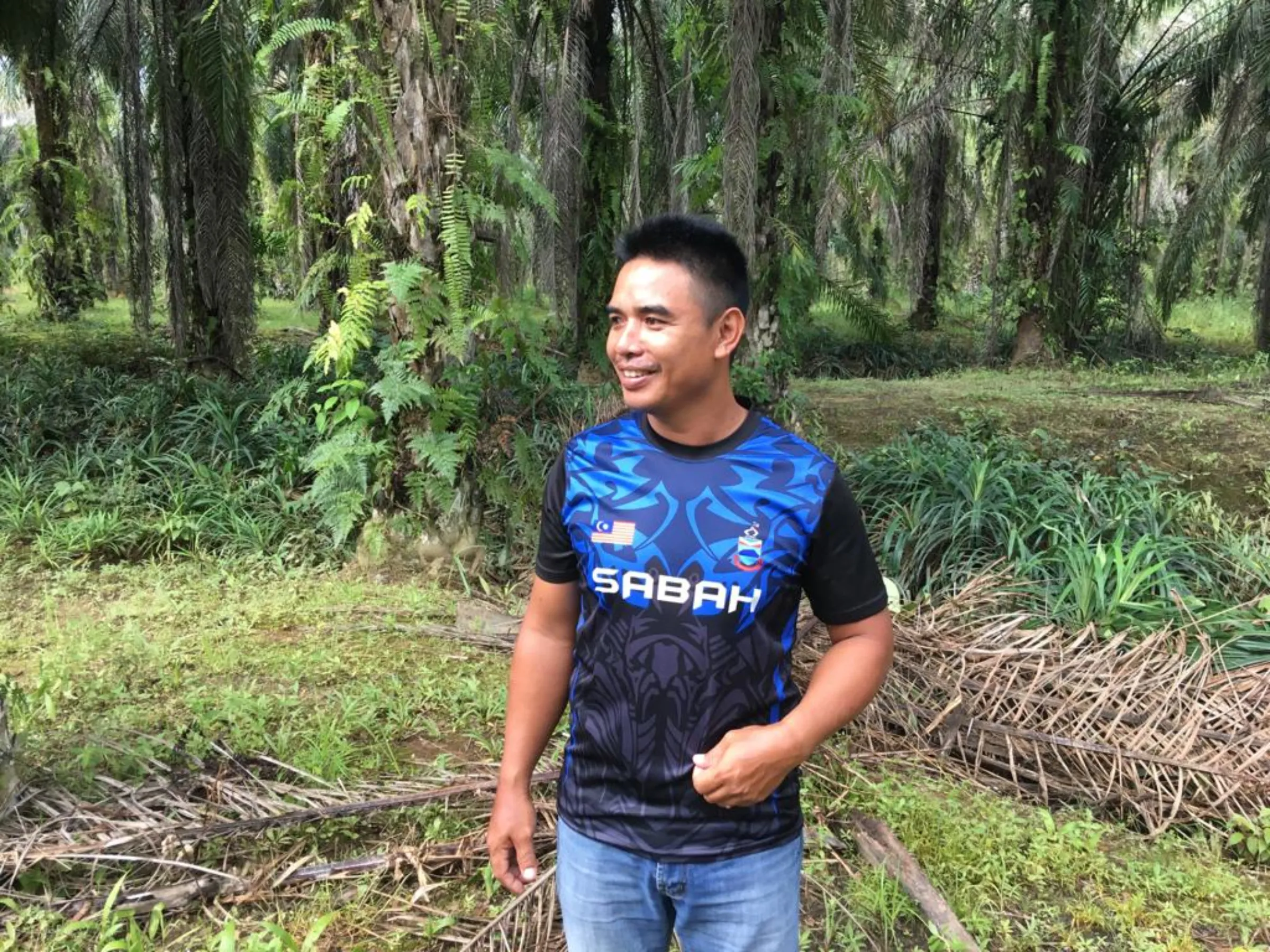 A man in a blue tshirt stands in a palm oil plantation