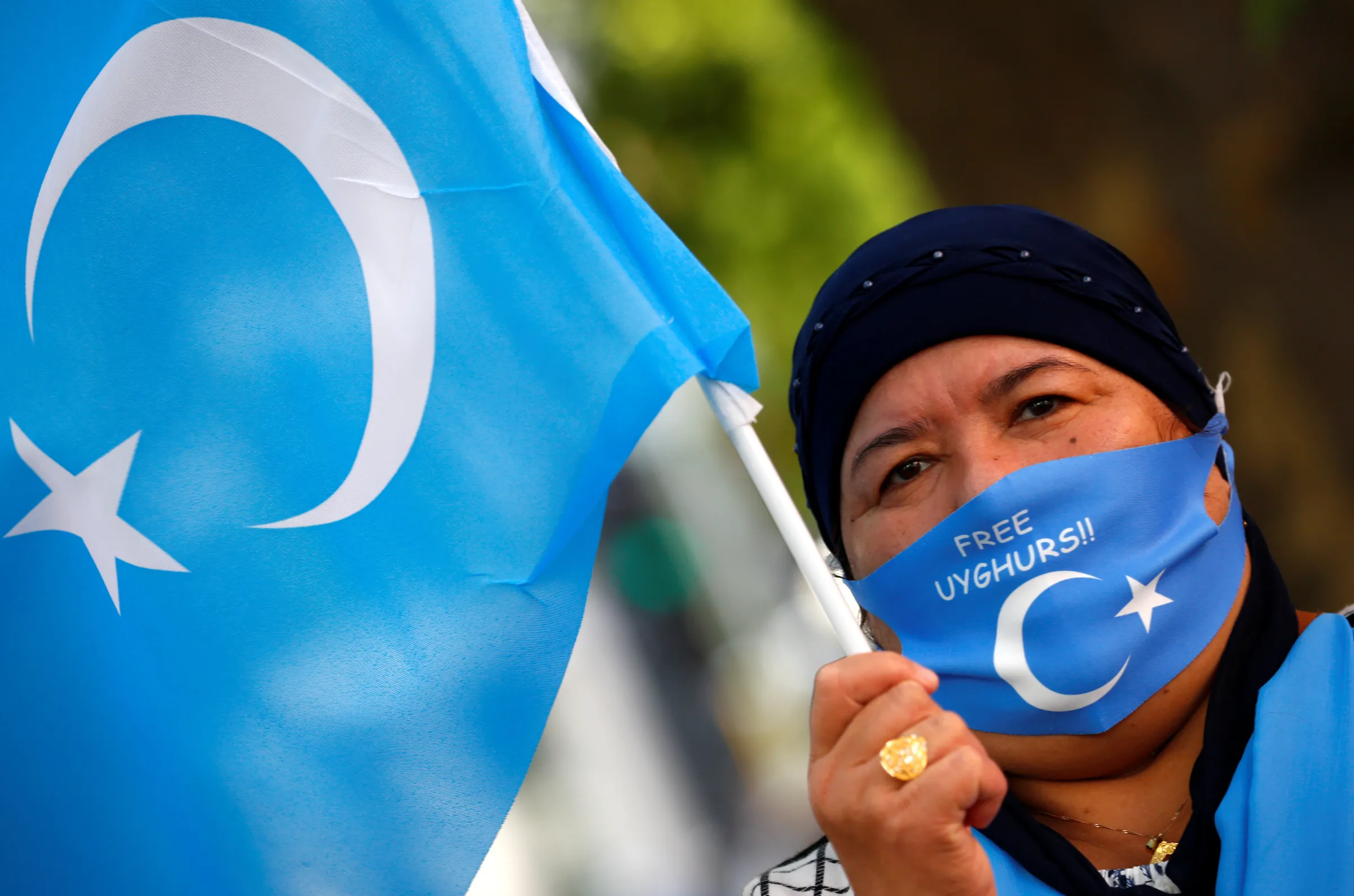 A woman holds a Uyghur flag and wears a Uyghur flag mask, where you can read 'FREE UYGHURS' at a rally
