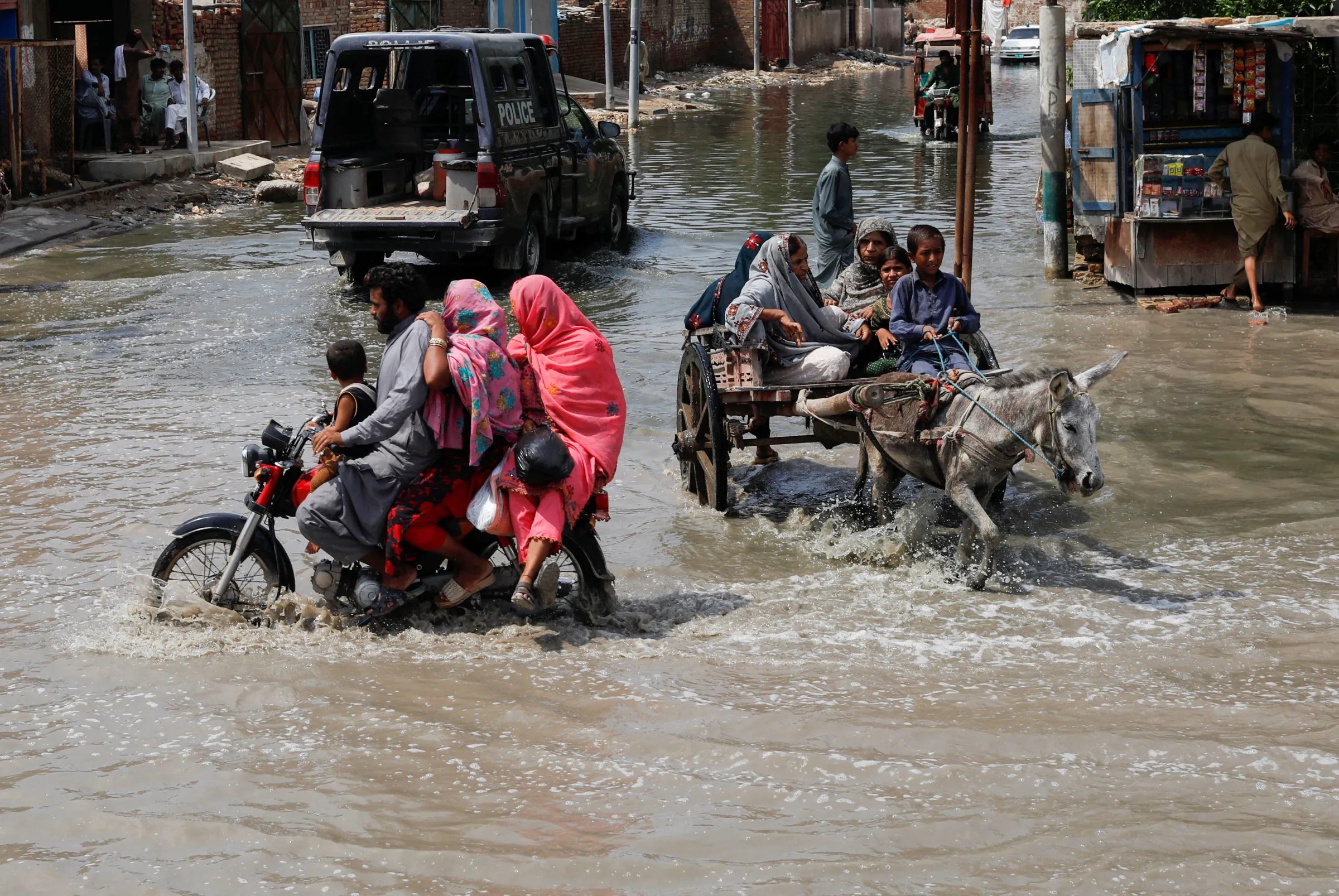 A family sit on a motorbike driving through floods next to a donkey pulling a cart with a family in