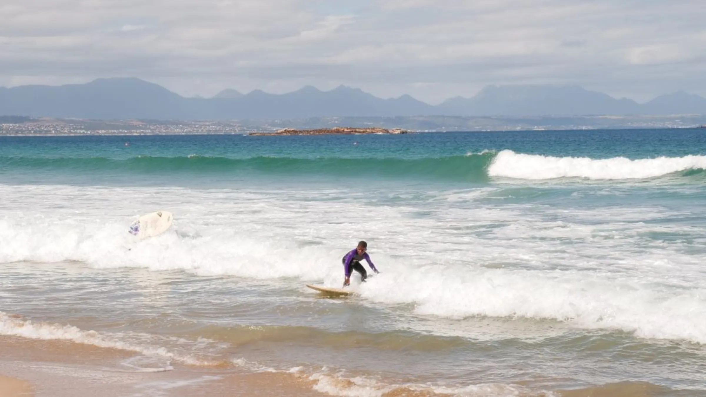 A student of The Surfer Kids charity catches a wave with the city and mountains seen behind him in the background in Mossel Bay, South Africa, November 25, 2022