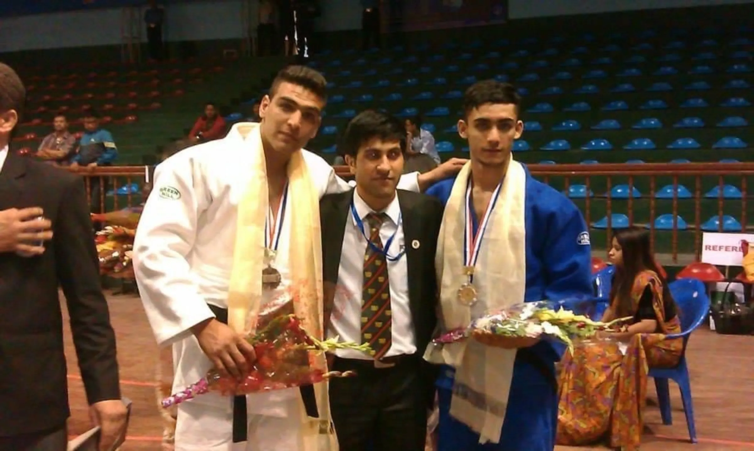 Three men are pictured side by side, two are in judogi and holding flowers